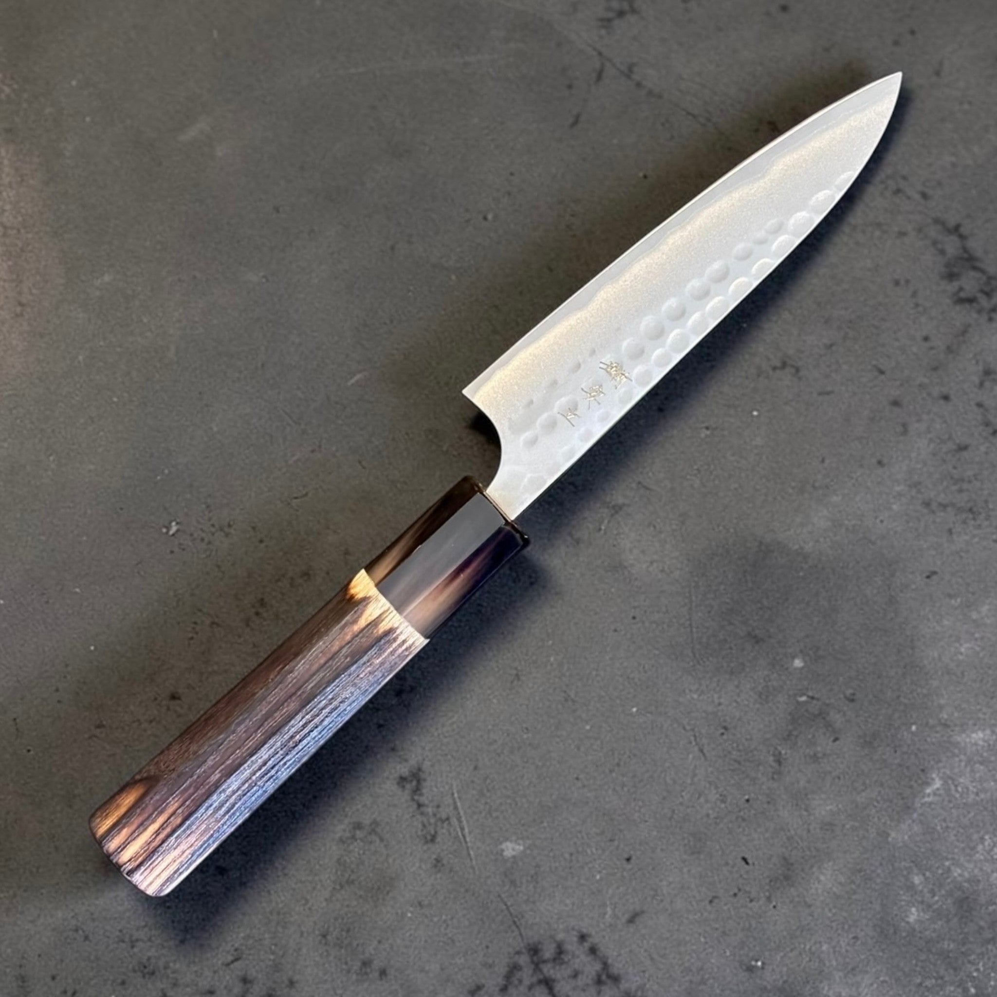 Anryu Aogami #2 Tsuchime Petty 120 mm with burnt wood handle and black buffalo horn ferrule, angled close-up showing hammered blade texture and burnt wood handle.