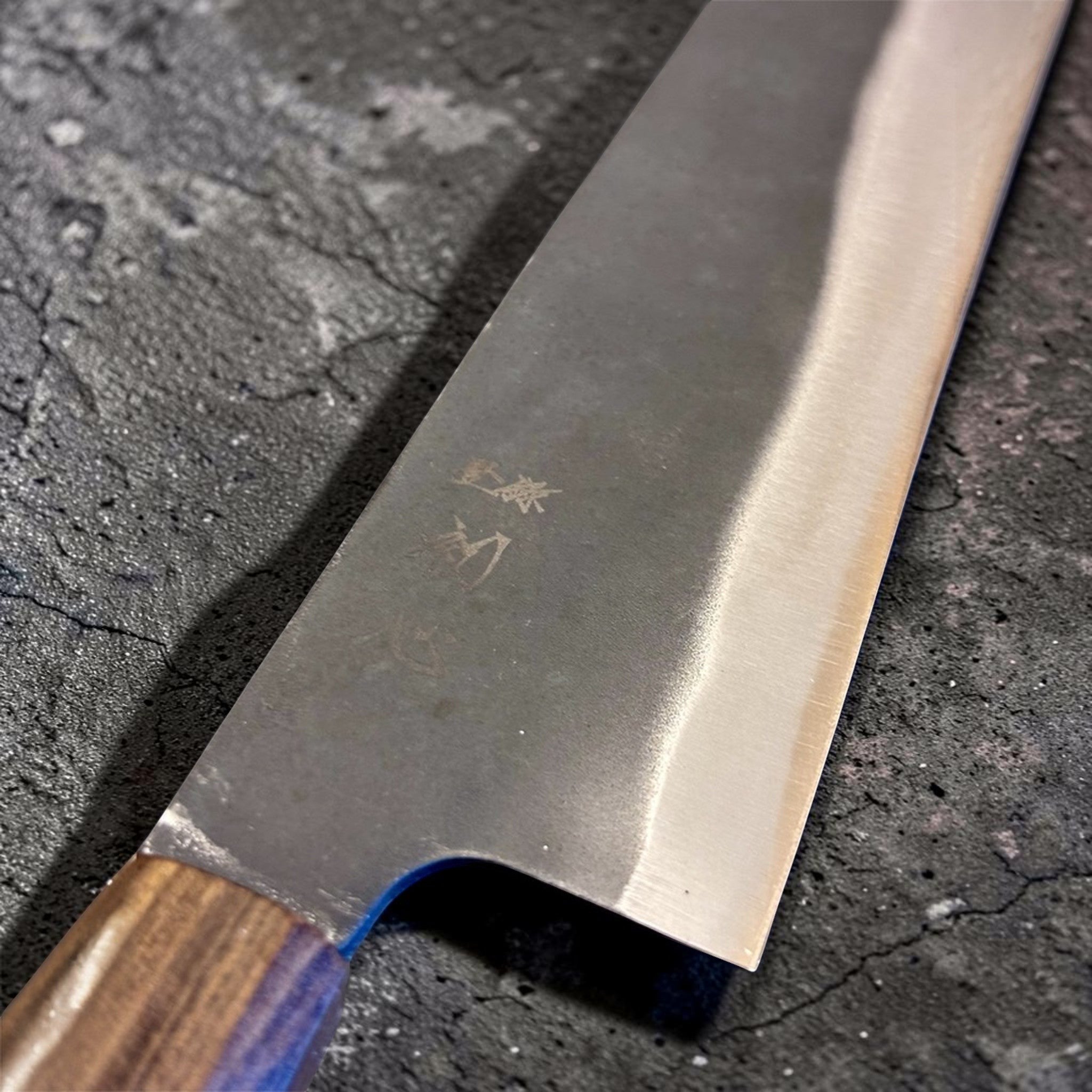 Close-up of the Hatsukokoro Blue #2 Kurouchi Bunka 190 mm blade near the heel, showing the engraved kanji, textured kurouchi cladding, and misty transition into the polished bevel above the edge.