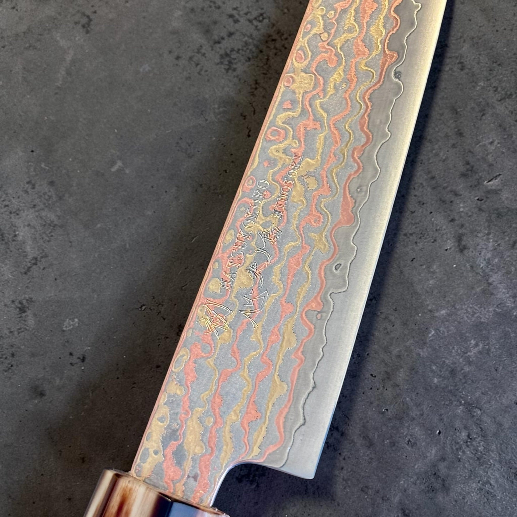 Close-up of the Hatsukokoro Hinotori V-Toku2 150mm petty blade showing the etched copper, brass, and steel rainbow Damascus pattern.