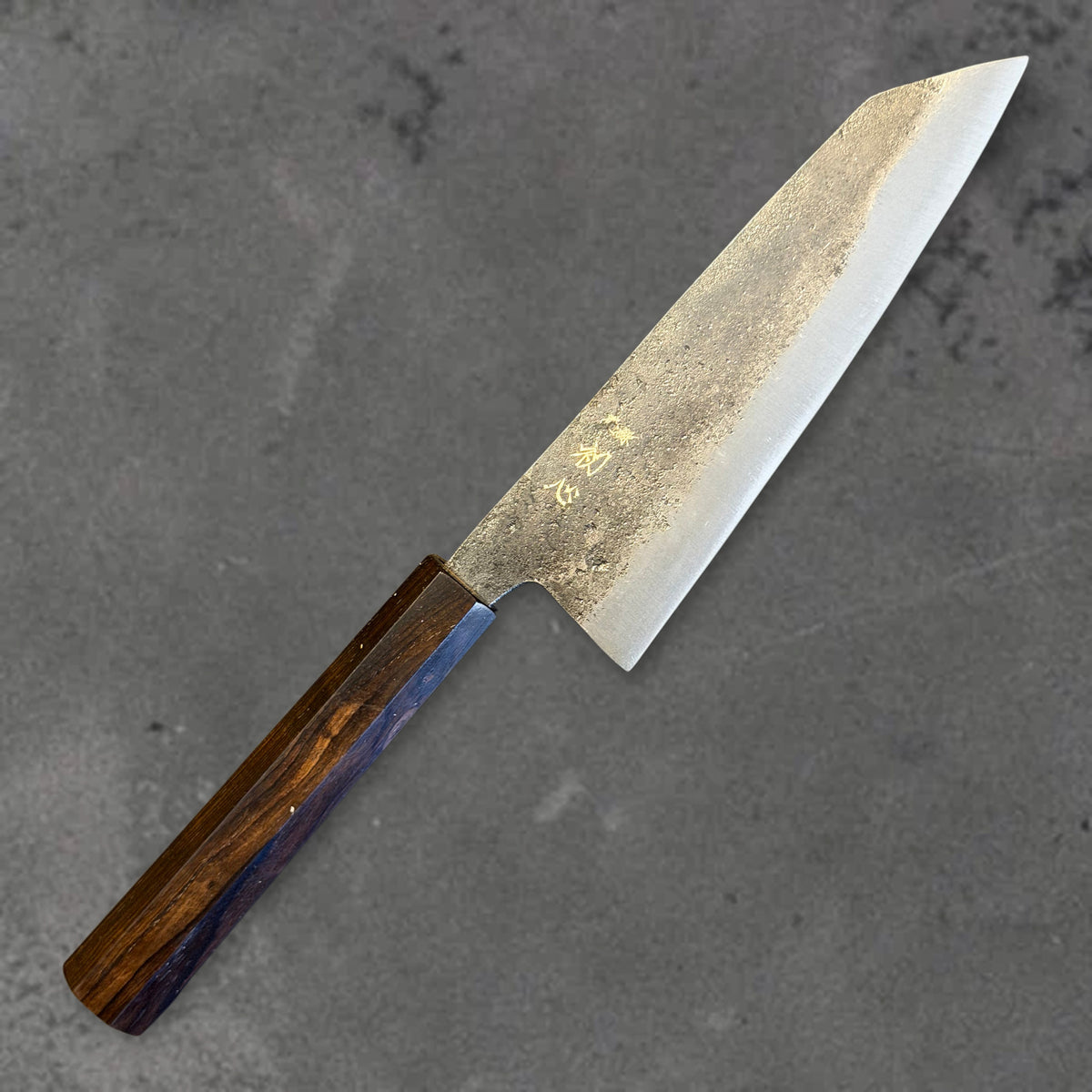 Angled front view of the Hatsukokoro Kurogane Blue #2 Kurouchi 140mm bunka with ebony handle, showing the blade shape, finish, and handle.