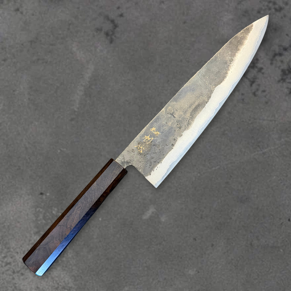 Angled front view of the Hatsukokoro Kurogane Blue #2 Kurouchi 210mm gyuto with ebony handle, showing the blade shape, finish, and handle.