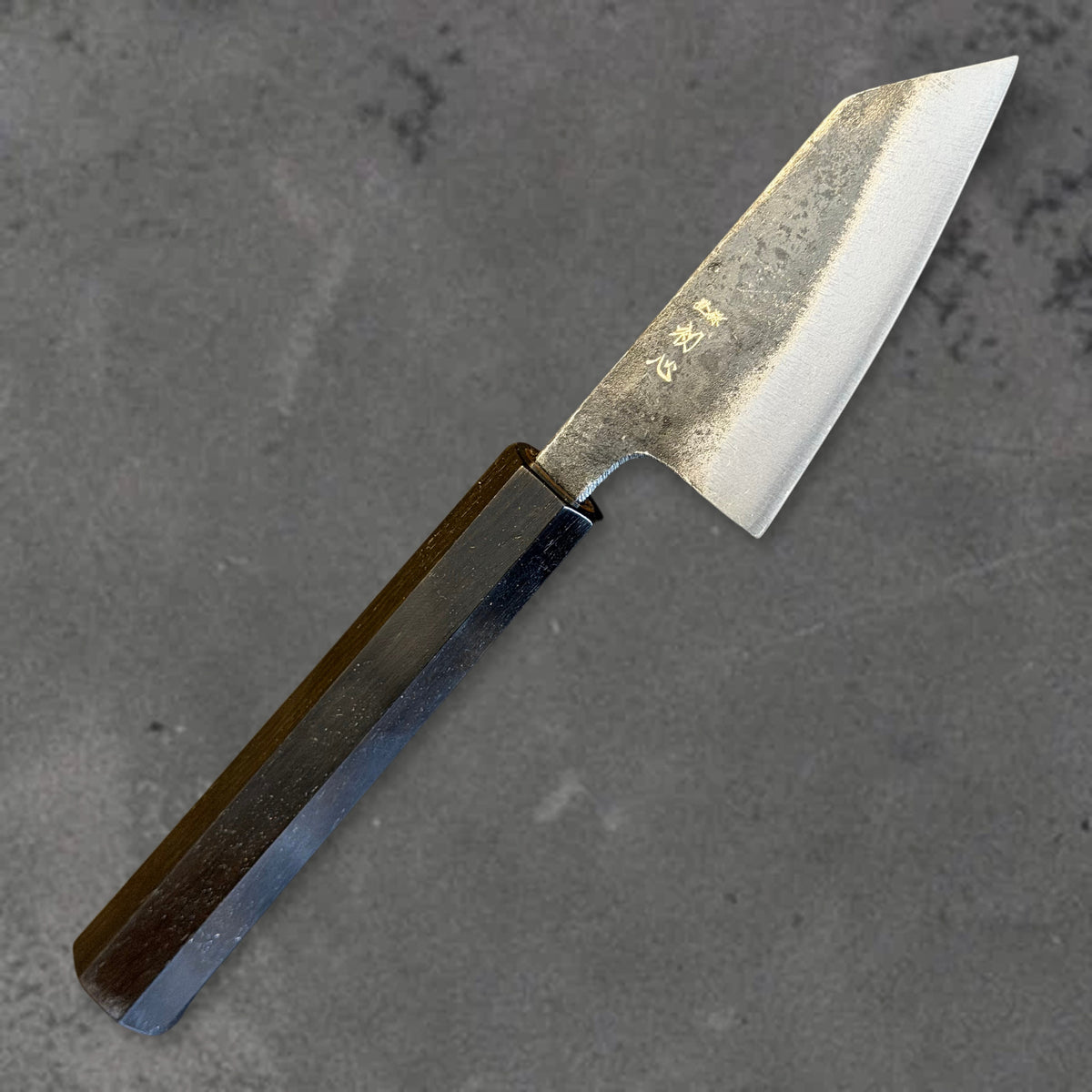 Angled front view of the Hatsukokoro Kurogane Blue #2 Kurouchi 80mm ko-bunka with ebony handle, showing the blade shape, finish, and handle.