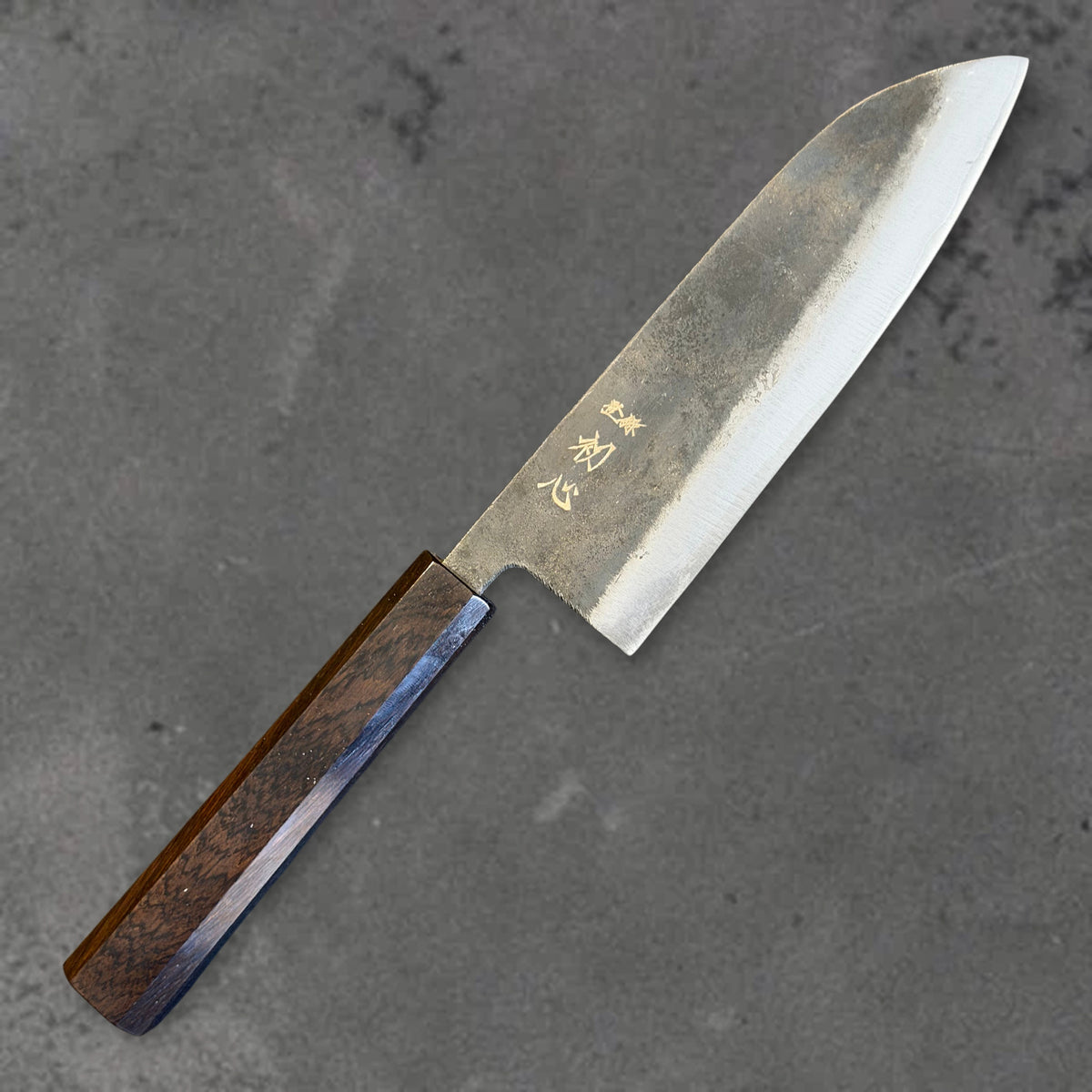 Angled front view of the Hatsukokoro Kurogane Blue #2 Kurouchi 165mm santoku with ebony handle, showing the blade shape, finish, and handle.