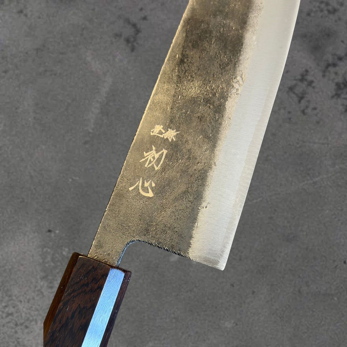 Close-up of the Kurouchi finish, blade markings, and polished bevel on the Hatsukokoro Kurogane Blue #2 Kurouchi 165mm santoku with ebony handle.