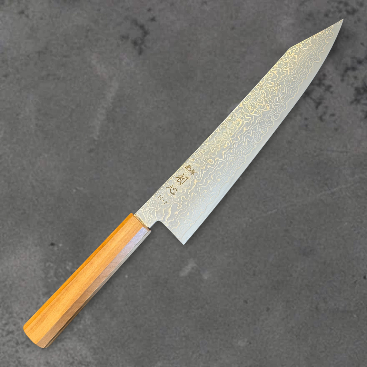 Angled front view of the Hatsukokoro Ryuhyo SG2 Damascus 210mm kiritsuke with olive wood handle, showing the blade shape, finish, and handle.