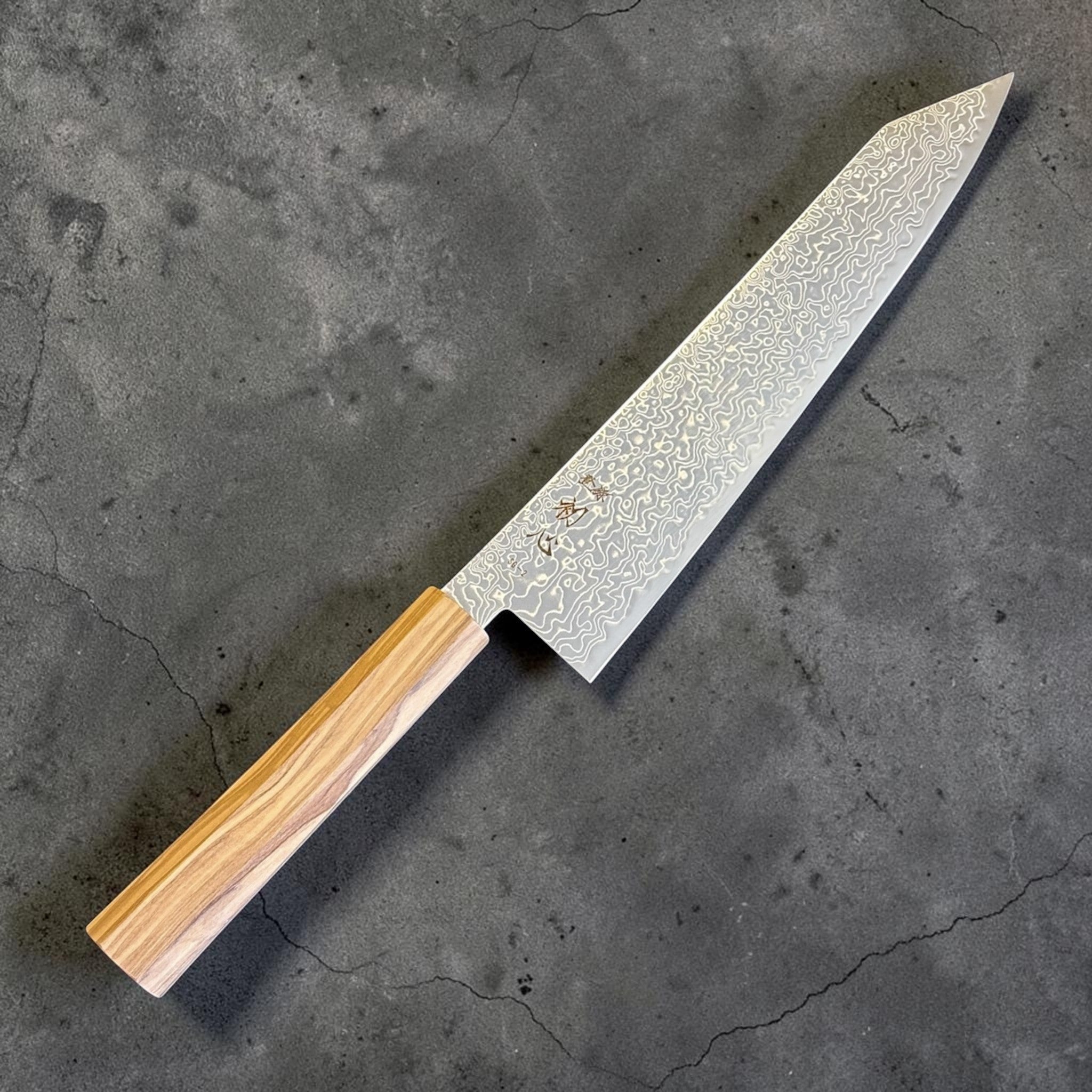 Hatsukokoro Ryuhyo SG2 Damascus Migaki Kiritsuke 240 mm with olive wood handle, angled full-length view showing the pointed kiritsuke tip and flowing Damascus pattern.