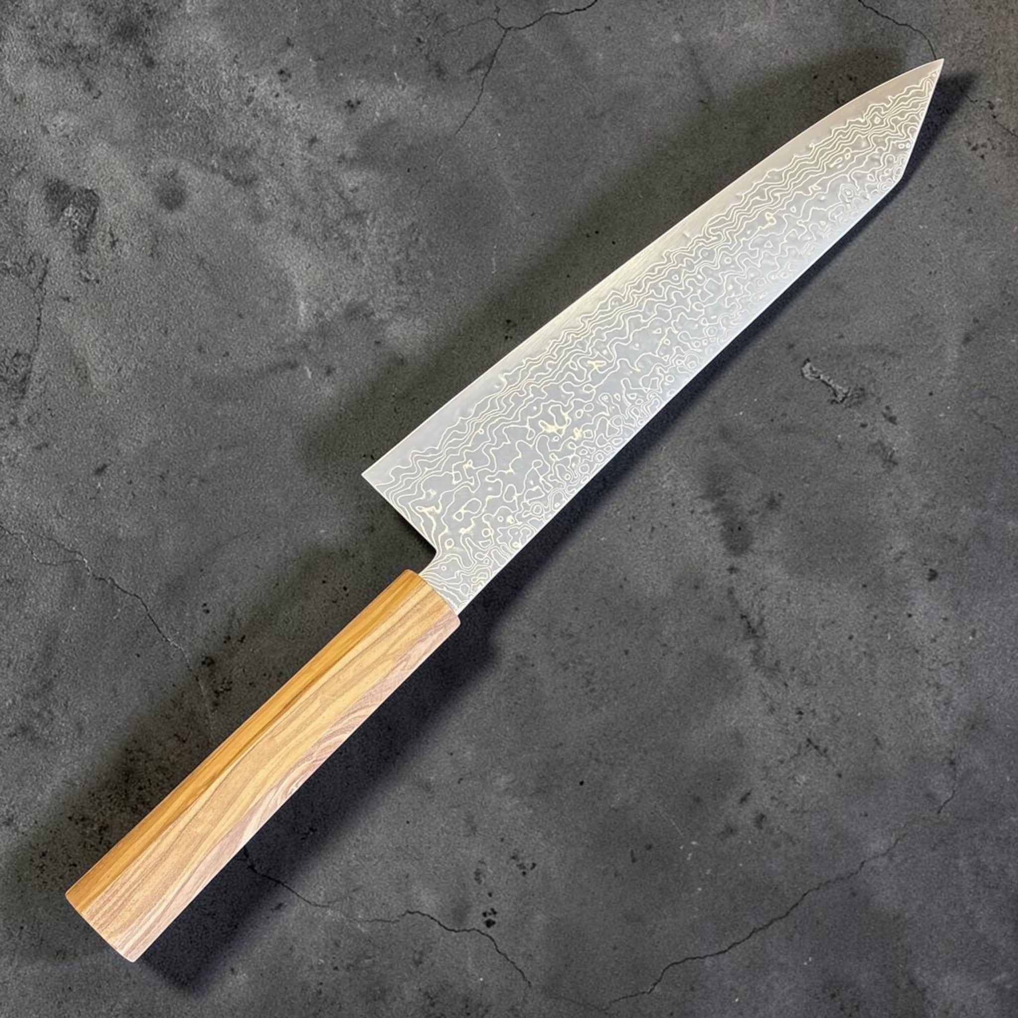 Hatsukokoro Ryuhyo SG2 Damascus Migaki Kiritsuke 240 mm with olive wood handle, diagonal blade-side view highlighting the long profile, polished migaki finish, and layered Damascus waves.