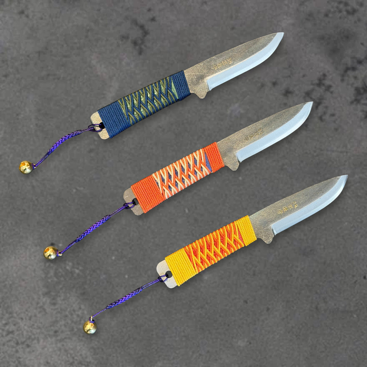 Three unsheathed Higonokami Bannou knives with blue, orange, and yellow wrapped handles, showing their blades and cord lanyards.