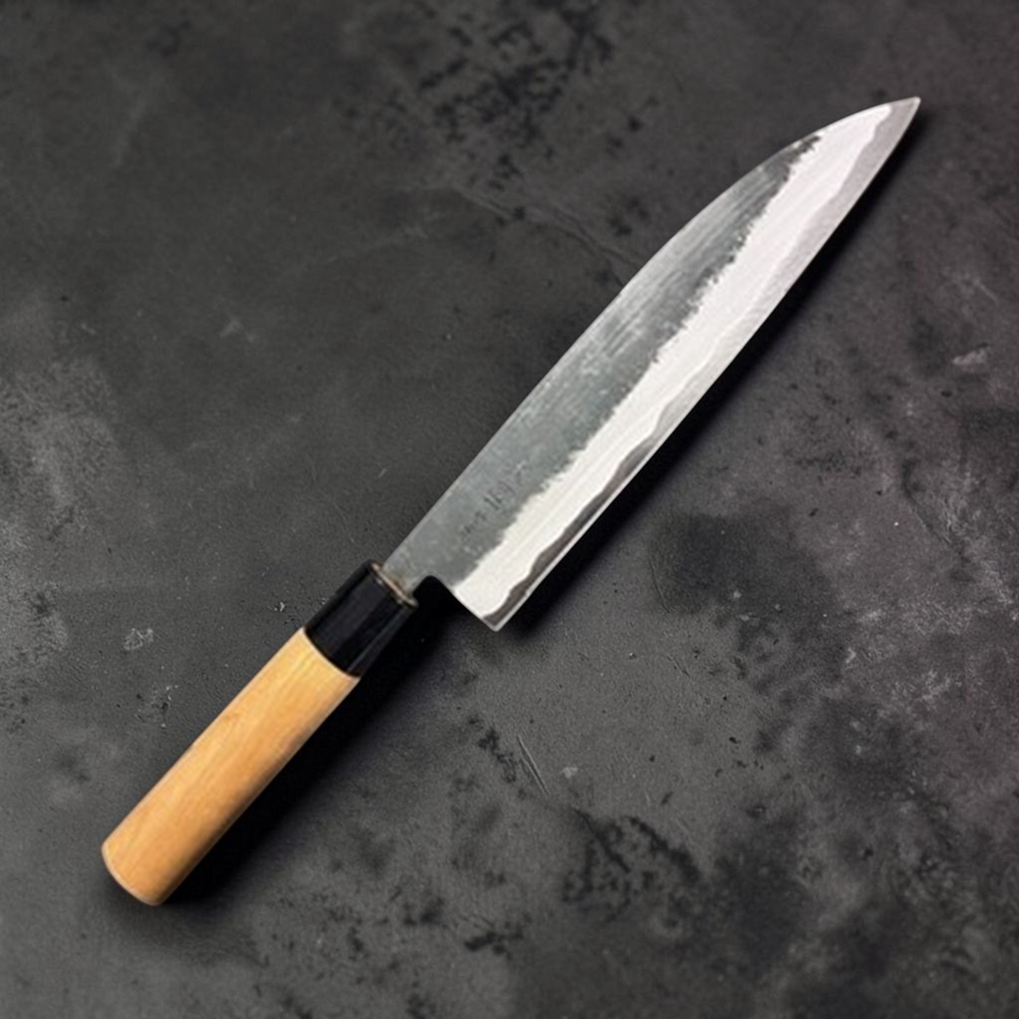 Hinokuni Aogami Super Kurouchi Gyuto 210 mm, three‑quarter view showing the kurouchi finish and light wa‑handle with black ferrule.
