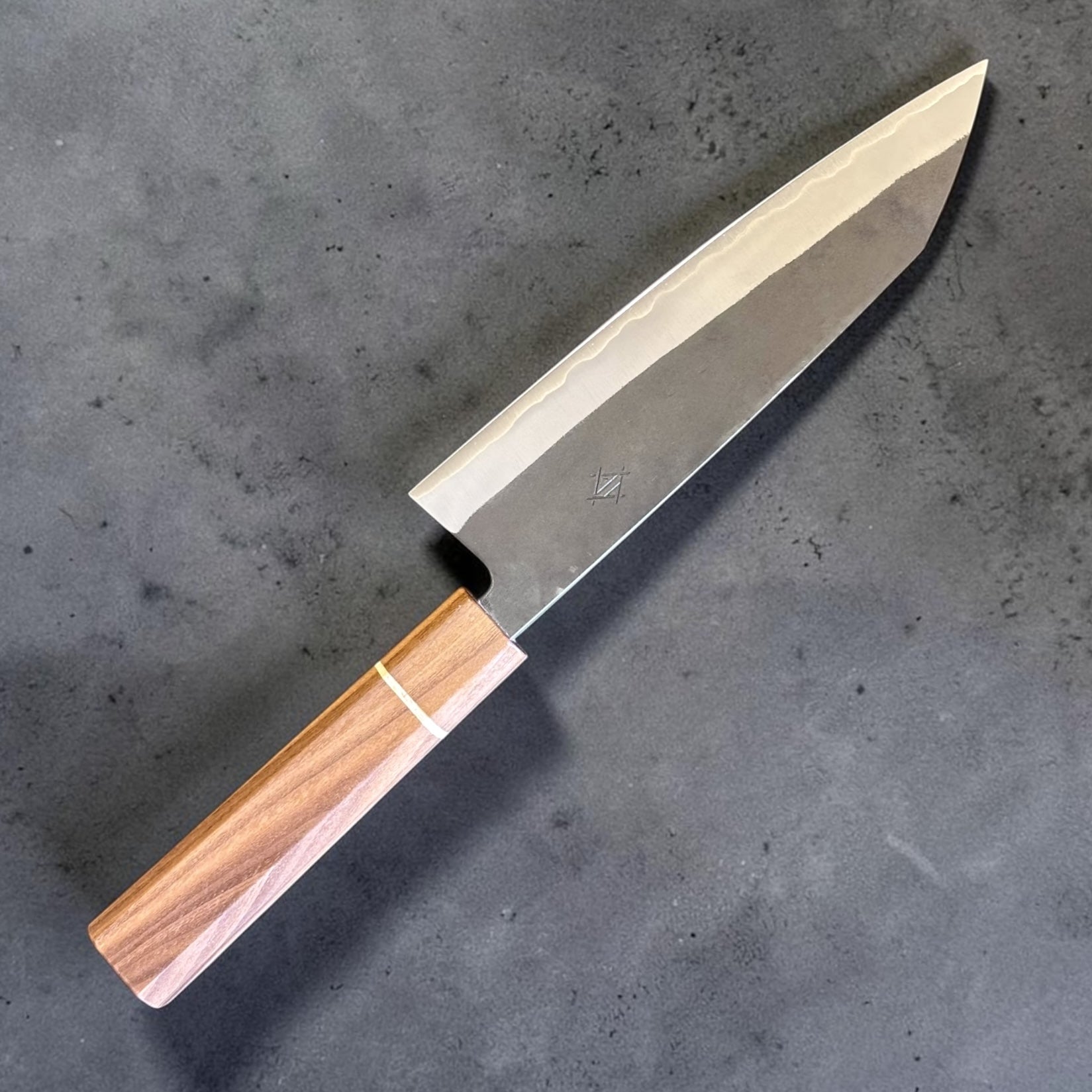 Three-quarter front view of the Imai Stainless Clad Aogami Super Kurouchi Bunka 183 mm showcasing the K-tip profile, kurouchi finish, and Sanjo walnut and maple stripe handle.