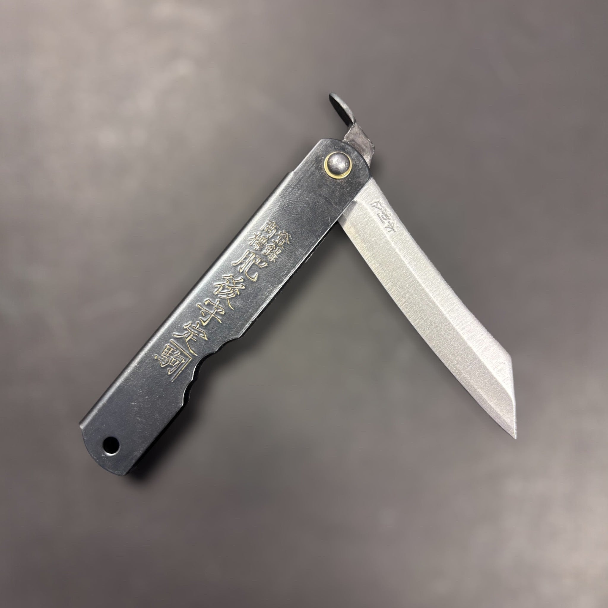 Nagao Higonokami SK carbon steel medium Japanese friction folder, black handle with gold kanji, blade partially open on dark grey background.