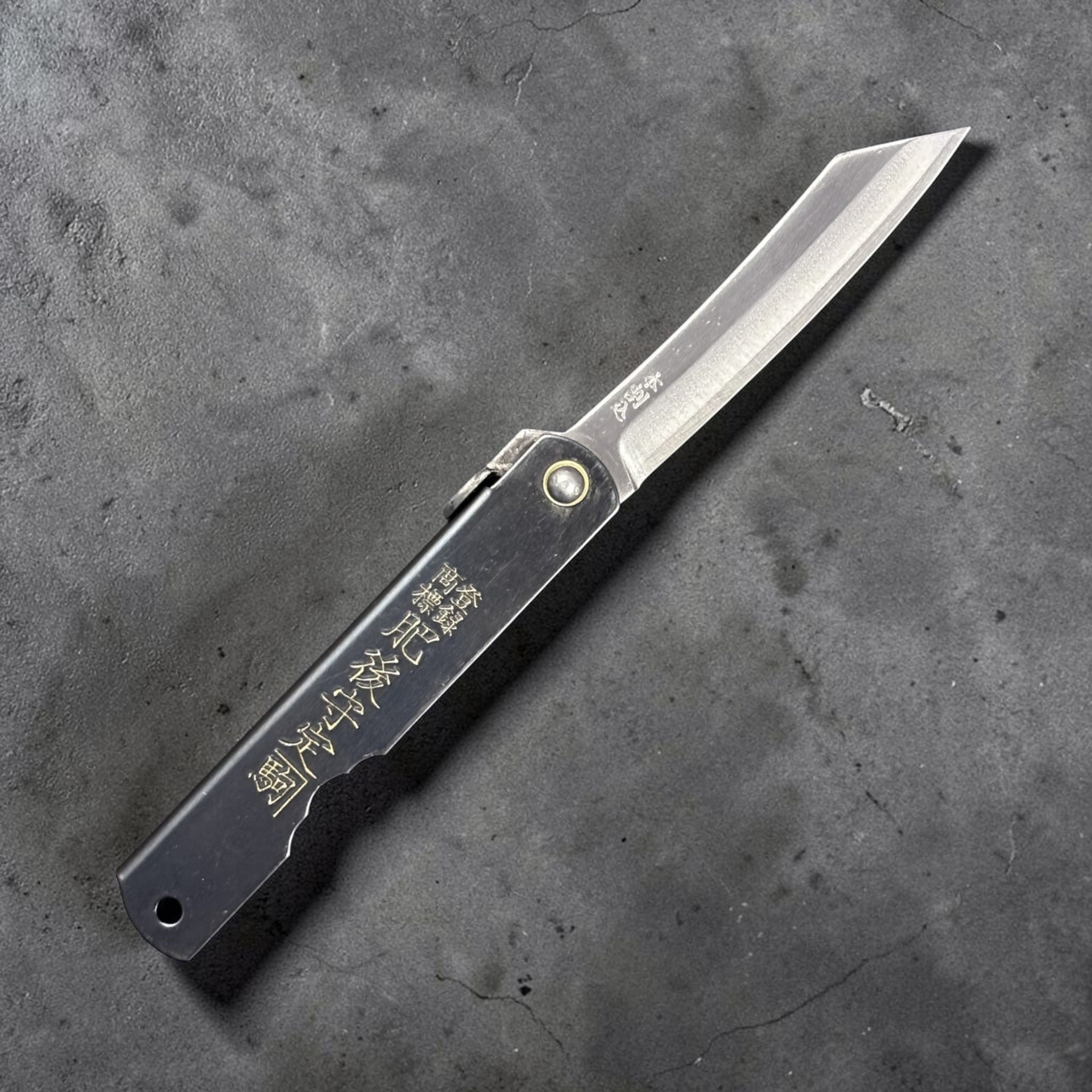 Nagao Higonokami SK carbon steel medium friction folder fully open, showing curved SK carbon blade and engraved black handle on neutral grey backdrop.