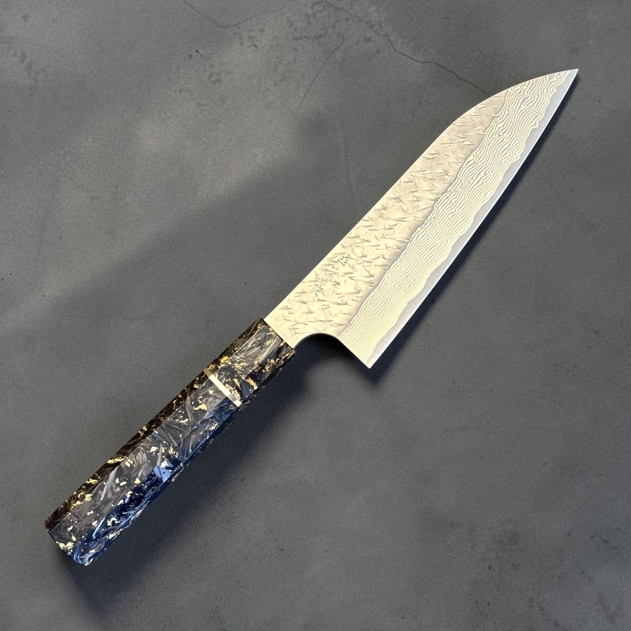 Angled view of the Nigara SG-STRIX Ko Santoku 150 mm highlighting the polished edge, tsuchime hammered finish, Damascus pattern and custom carbon fiber wa handle.