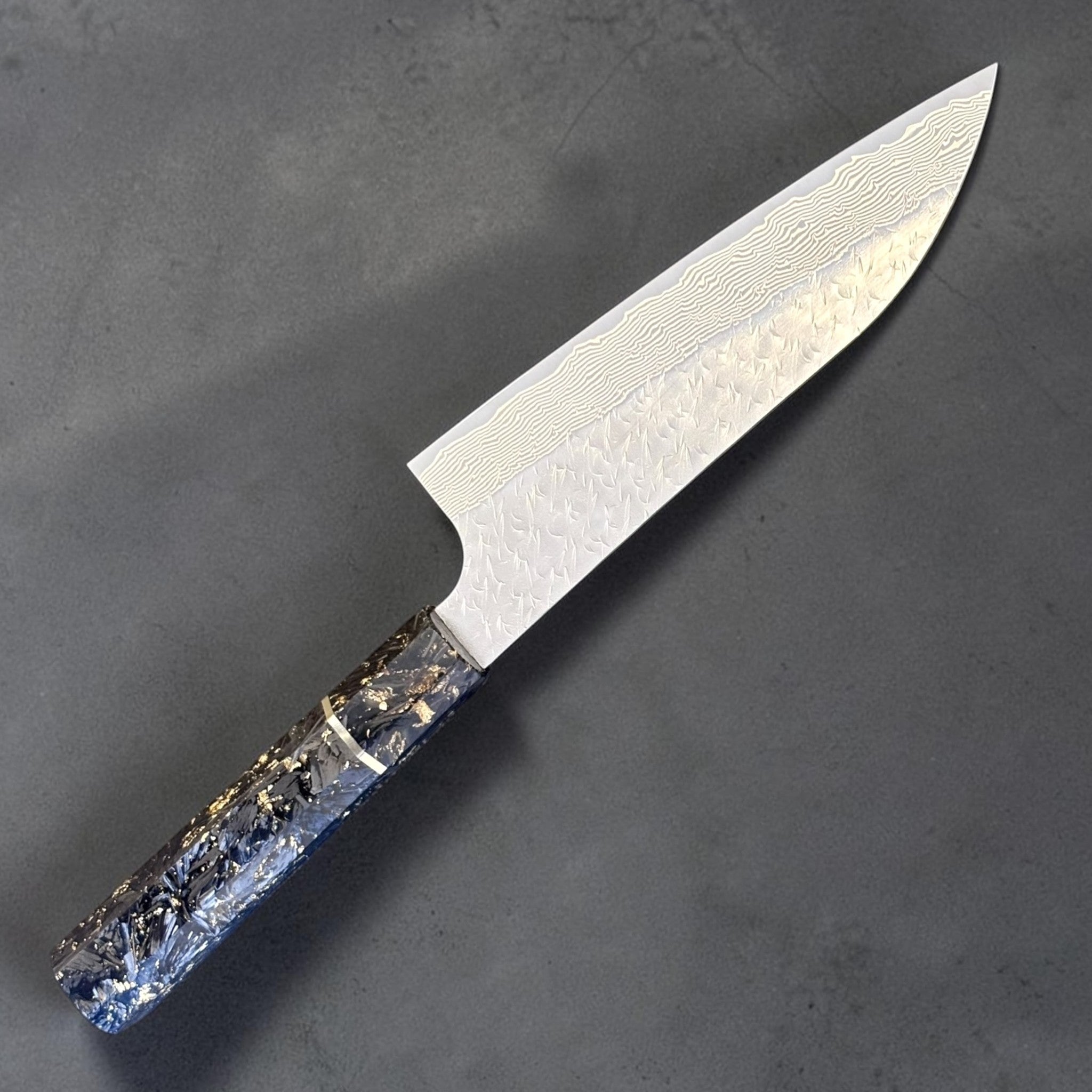 Three-quarter view of the Nigara SG-STRIX Ko Santoku 150 mm showcasing the flowing Damascus layers, hammered blade finish and premium carbon fiber handle with gold flecks.