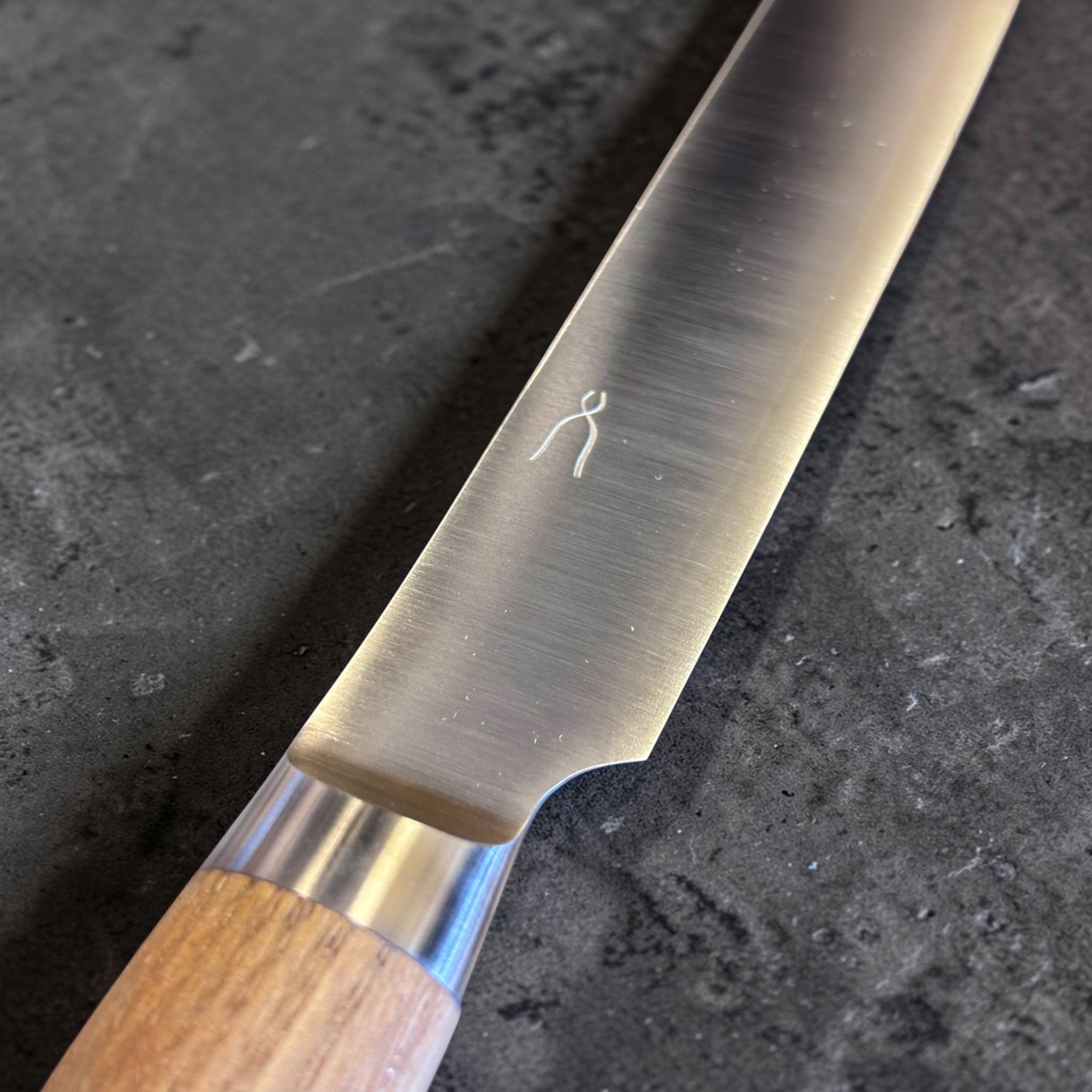 Tadafusa SLD Bread Knife with light wood handle, close-up of the blade face, maker mark, and bolster area.
