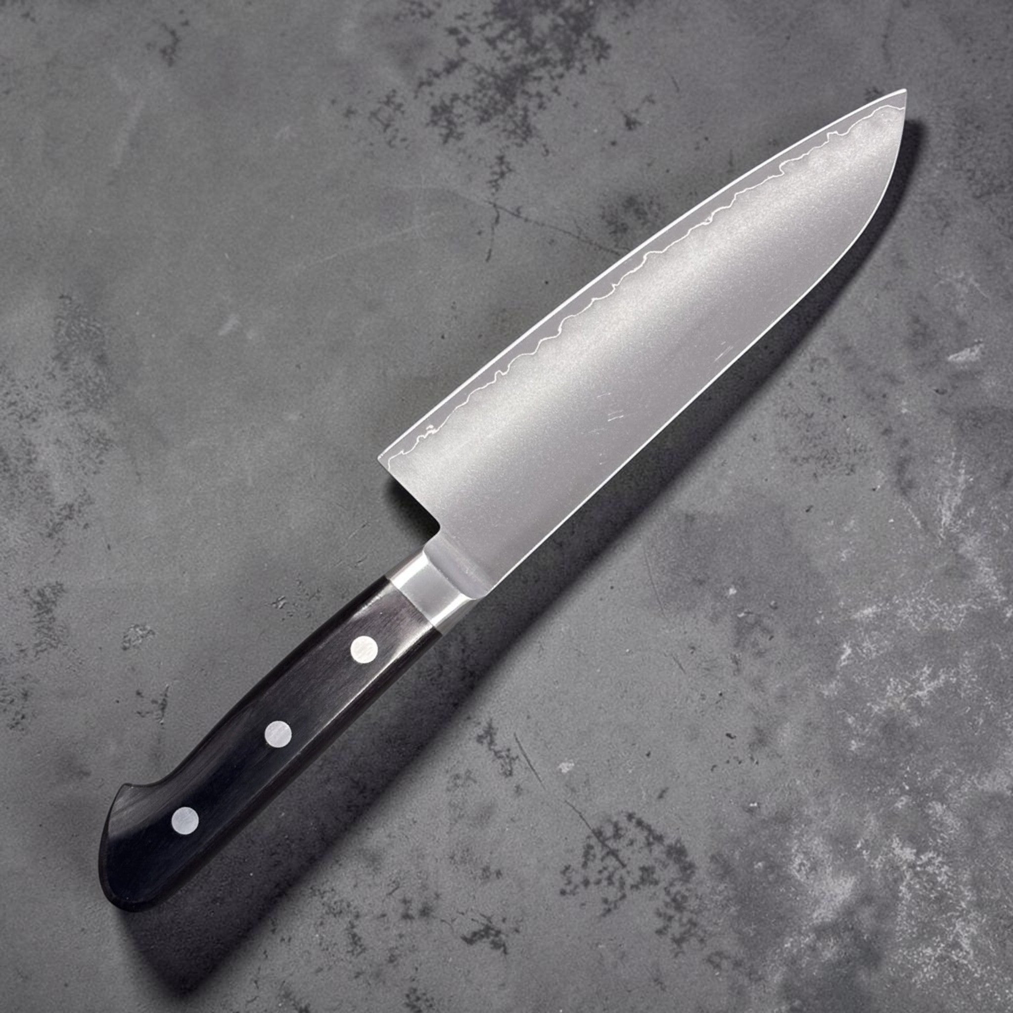Tsunehisa VG-1 stainless steel Santoku 165 mm with black Western handle, left side blade face highlighting the soft cladding line.