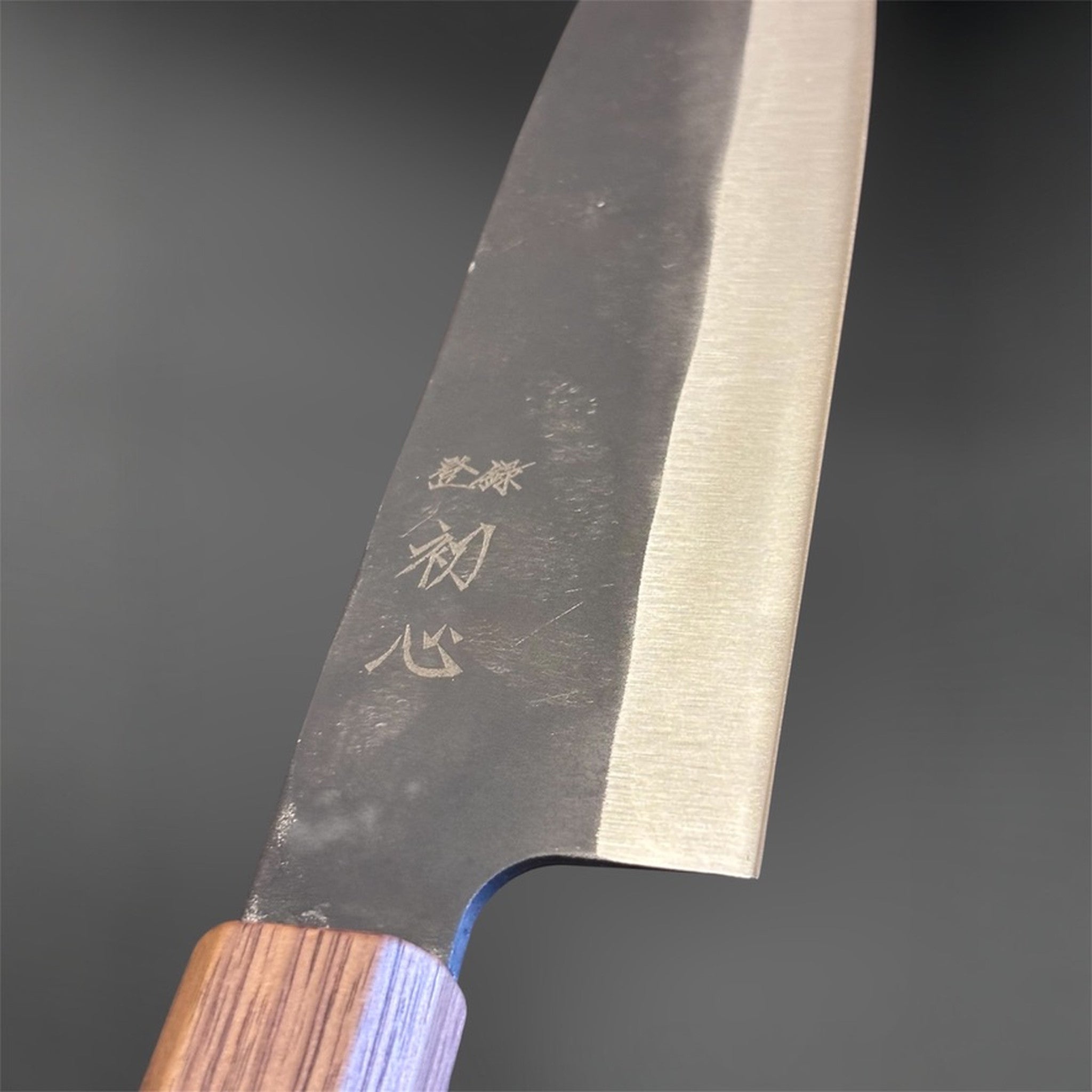 Close-up of the Hatsukokoro Aokitae Blue #2 Kurouchi Santoku 180 mm choil area, showing the kurouchi finish, engraved kanji, and Sanjo walnut handle with maple stripe.