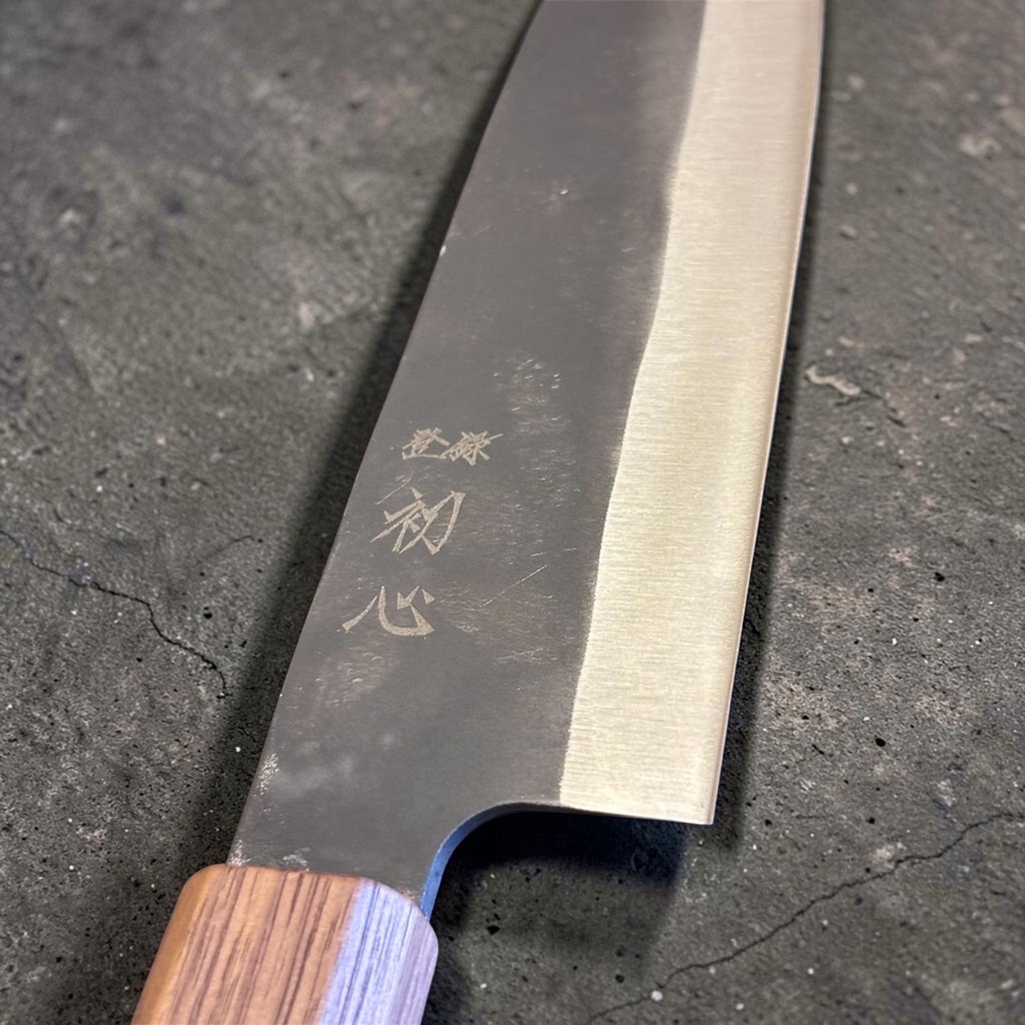 Close-up of the Hatsukokoro Aokitae Blue #2 Kurouchi Santoku 180 mm choil area, showing the kurouchi finish, engraved kanji, and Sanjo walnut handle with maple stripe.
