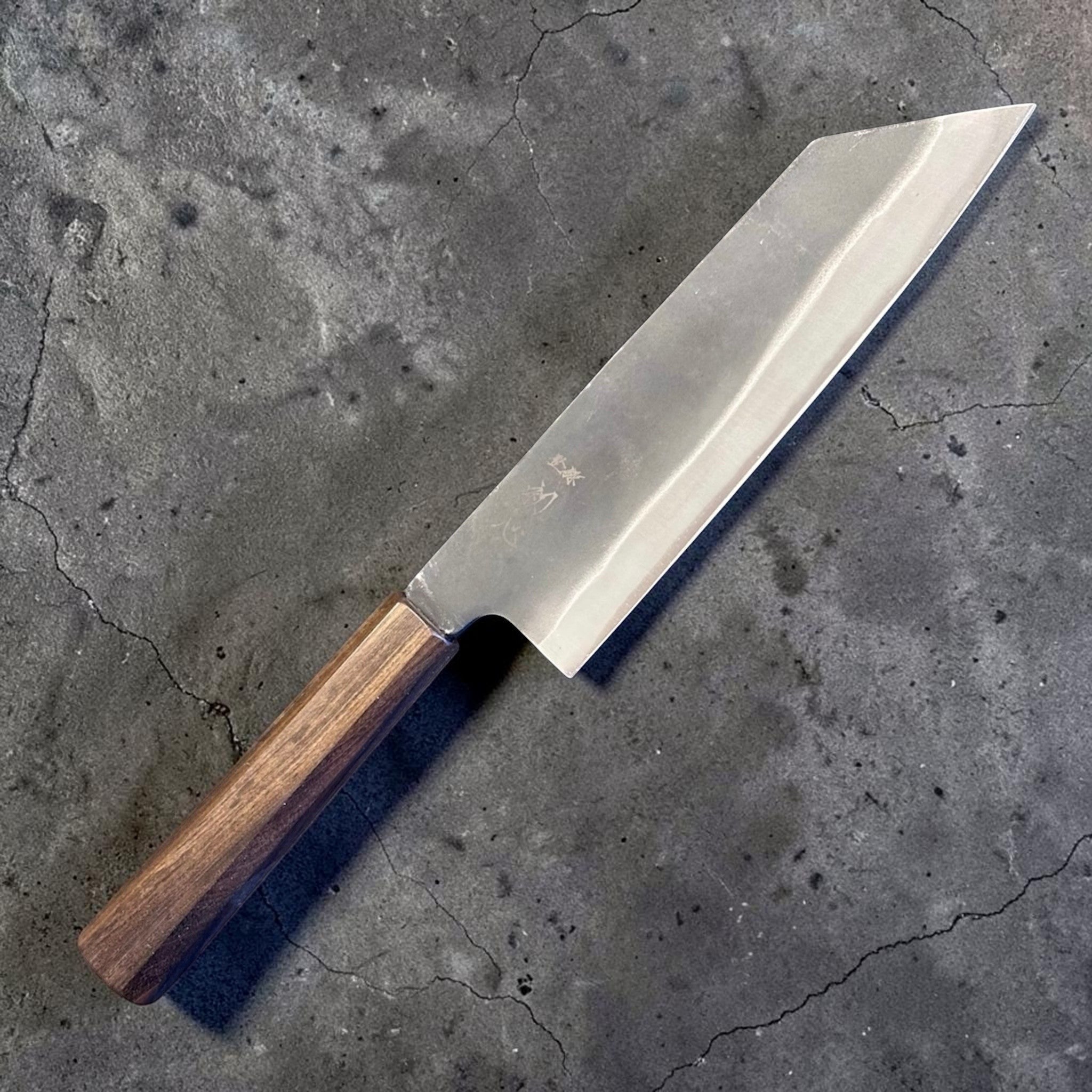 Hatsukokoro Blue #2 Kurouchi Bunka 190 mm with mono ebony handle, angled front-side product photo showing the dark kurouchi finish, wide bevel, and aggressive k-tip bunka profile.