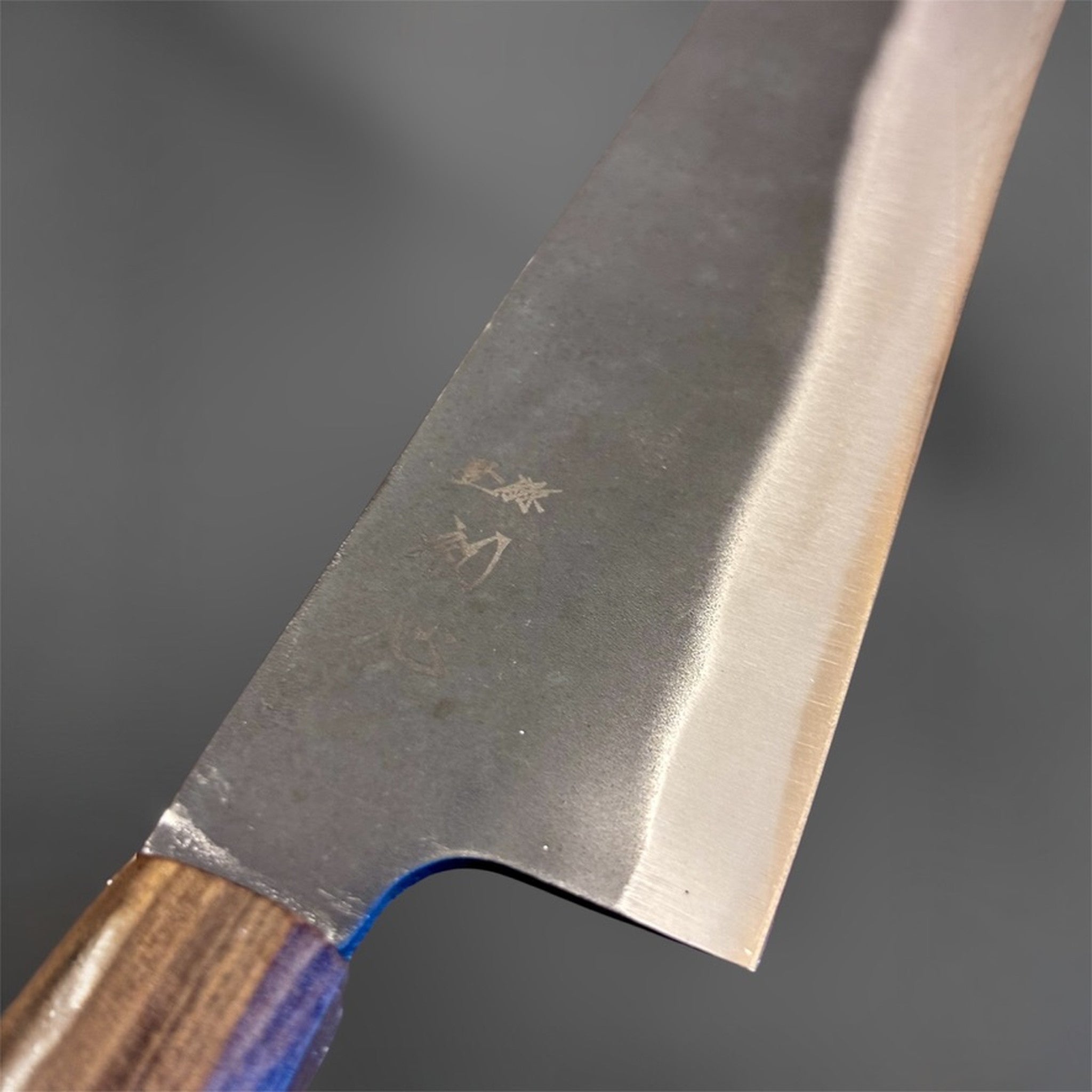 Close-up of the Hatsukokoro Blue #2 Kurouchi Bunka 190 mm blade near the heel, showing the engraved kanji, textured kurouchi cladding, and misty transition into the polished bevel above the edge.