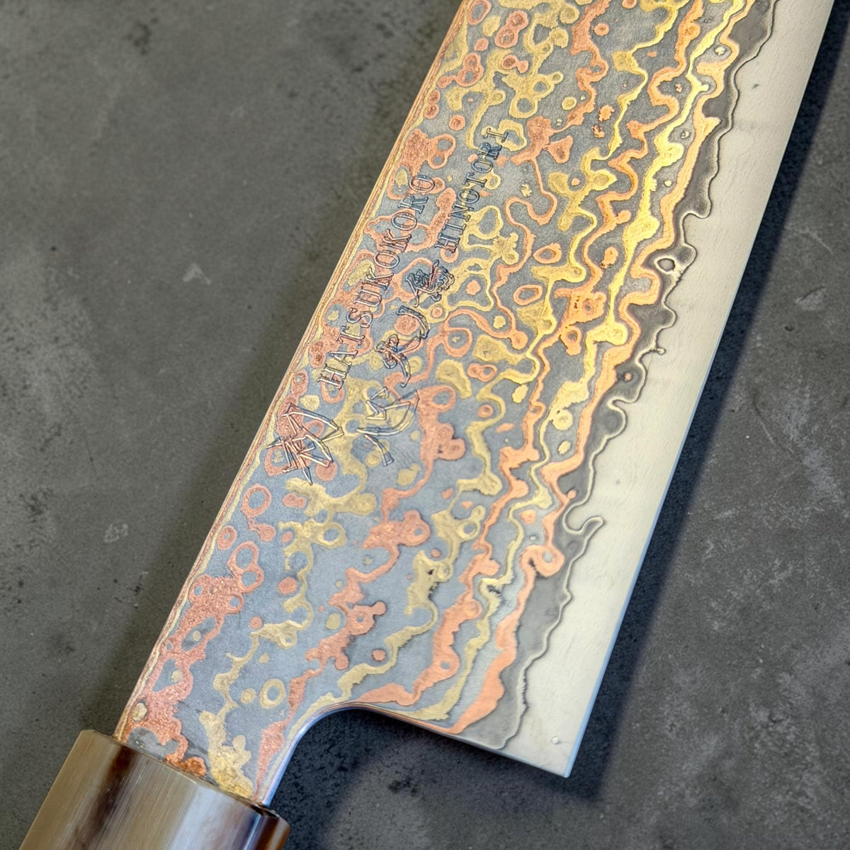 Hatsukokoro Hinotori Gyuto blade close-up showing V-Toku2 core, copper brass rainbow Damascus and kanji