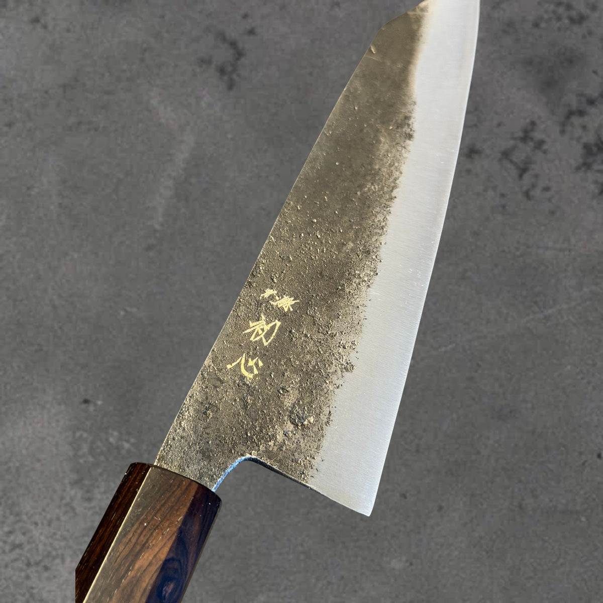 Close-up of the Kurouchi finish, blade markings, and polished bevel on the Hatsukokoro Kurogane Blue #2 Kurouchi 140mm bunka with ebony handle.