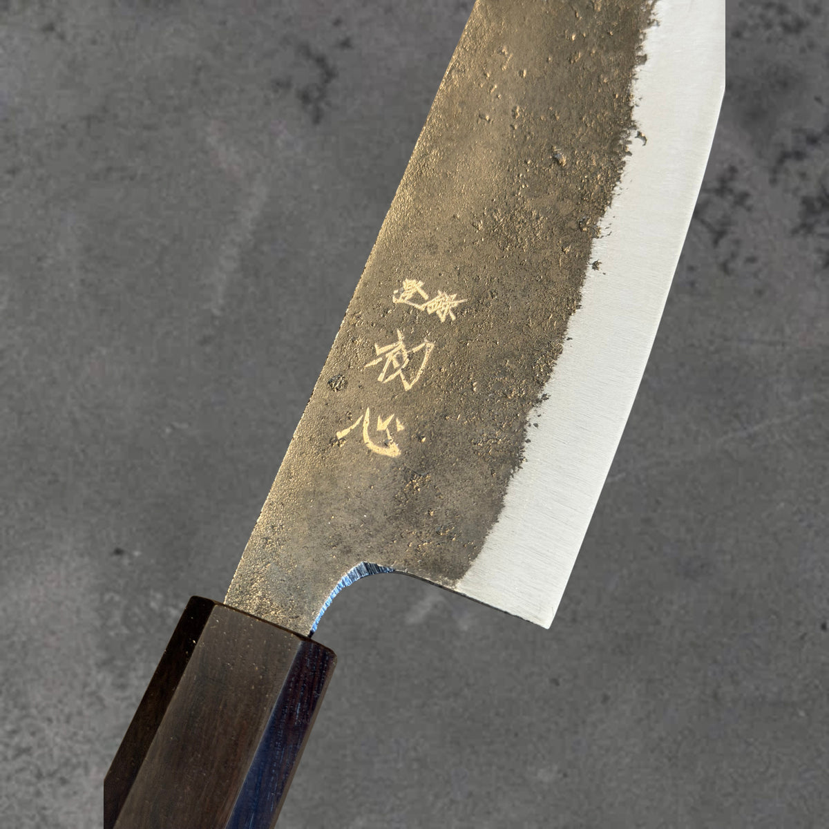Close-up of the Kurouchi finish, blade markings, and polished bevel on the Hatsukokoro Kurogane Blue #2 Kurouchi 165mm bunka with ebony handle.