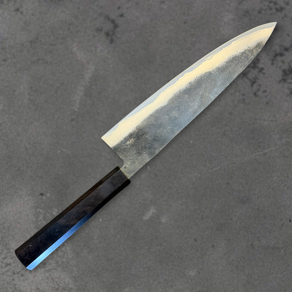 Angled reverse view of the Hatsukokoro Kurogane Blue #2 Kurouchi 210mm gyuto with ebony handle, showing the blade shape, finish, and handle.