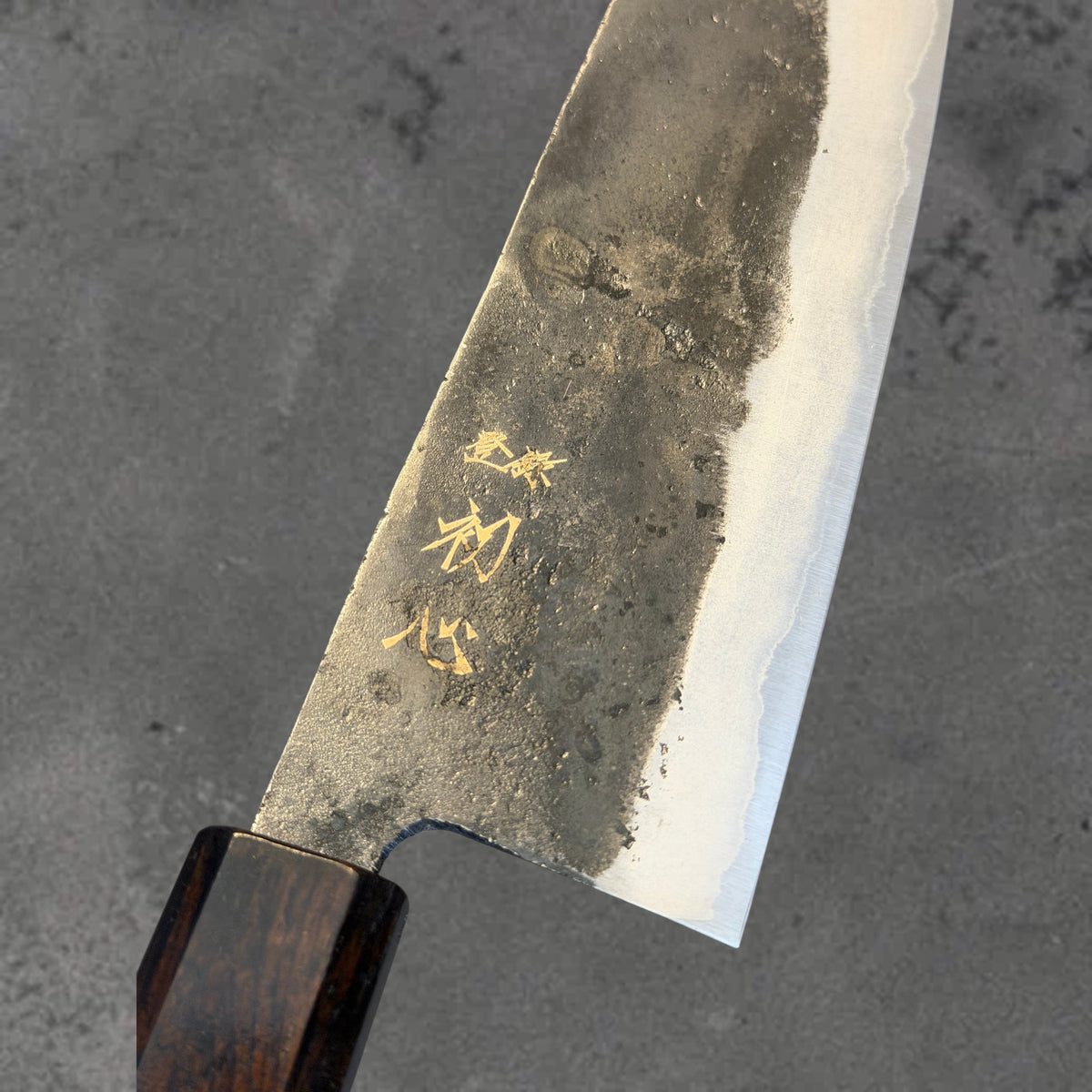 Close-up of the Kurouchi finish, blade markings, and polished bevel on the Hatsukokoro Kurogane Blue #2 Kurouchi 210mm gyuto with ebony handle.