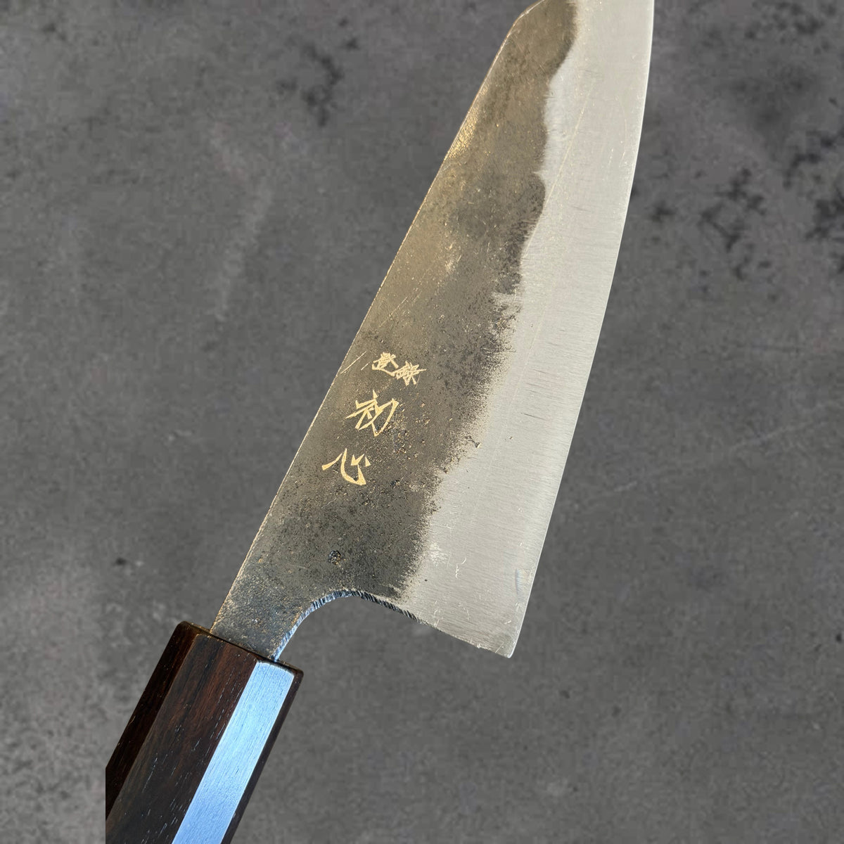 Close-up of the Kurouchi finish, blade markings, and polished bevel on the Hatsukokoro Kurogane Blue #2 Kurouchi 120mm ko-bunka with ebony handle.