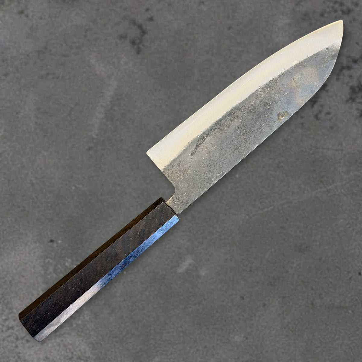 Angled reverse view of the Hatsukokoro Kurogane Blue #2 Kurouchi 165mm santoku with ebony handle, showing the blade shape, finish, and handle.