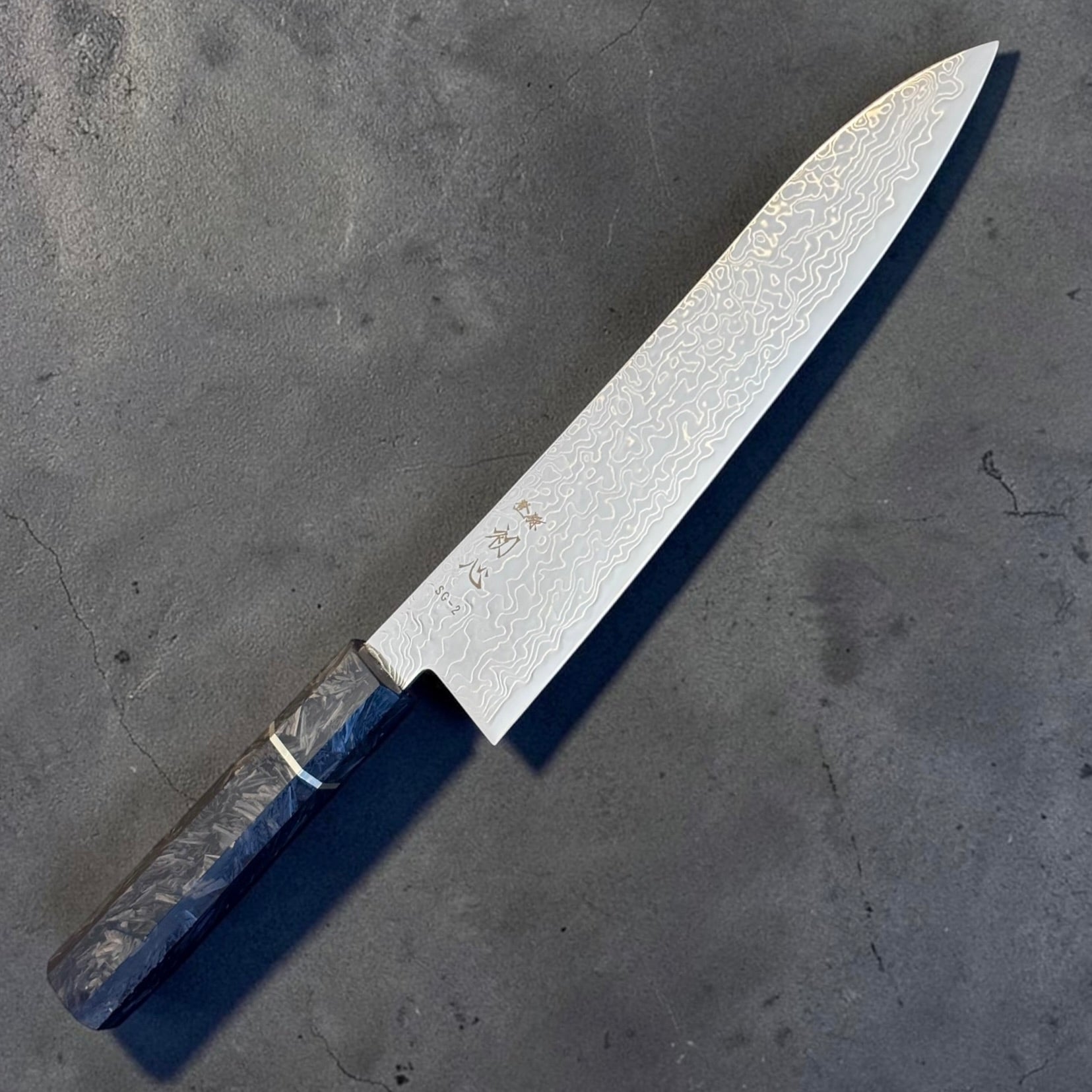 Reverse angled view of the Hatsukokoro Ryuhyo SG2 Damascus Gyuto 210mm and carbon fiber handle.