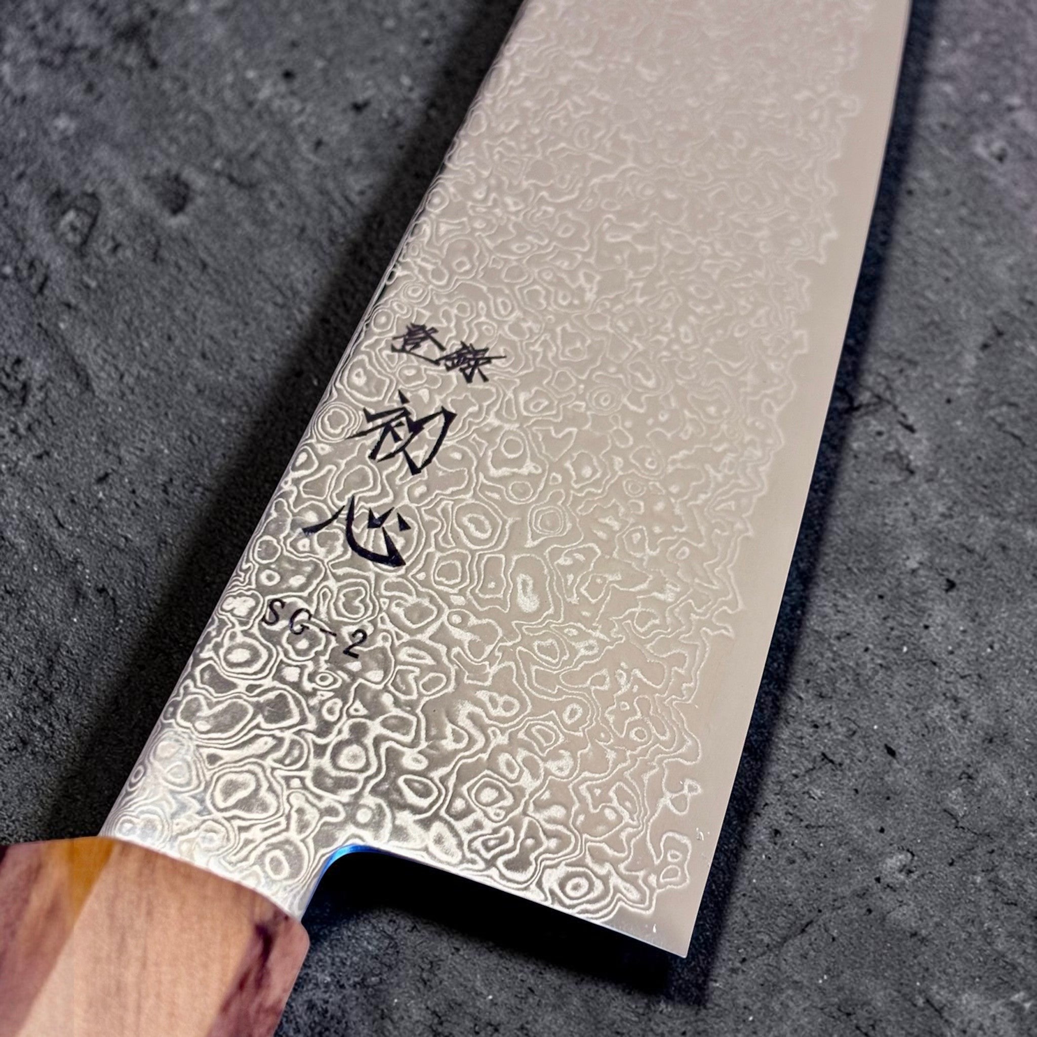 Hatsukokoro Saihyo SG2 Damascus Bunka 180 mm, close-up of the SG2 Damascus pattern, kanji engraving, and olivewood handle junction.