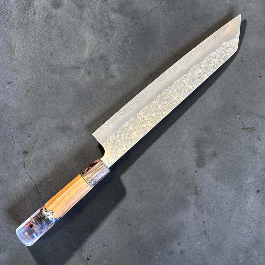Close-up of Makoto Kurosaki SG2 Bunka 240mm showing kasumi polish, highlighted cladding line and patterned hammered finish