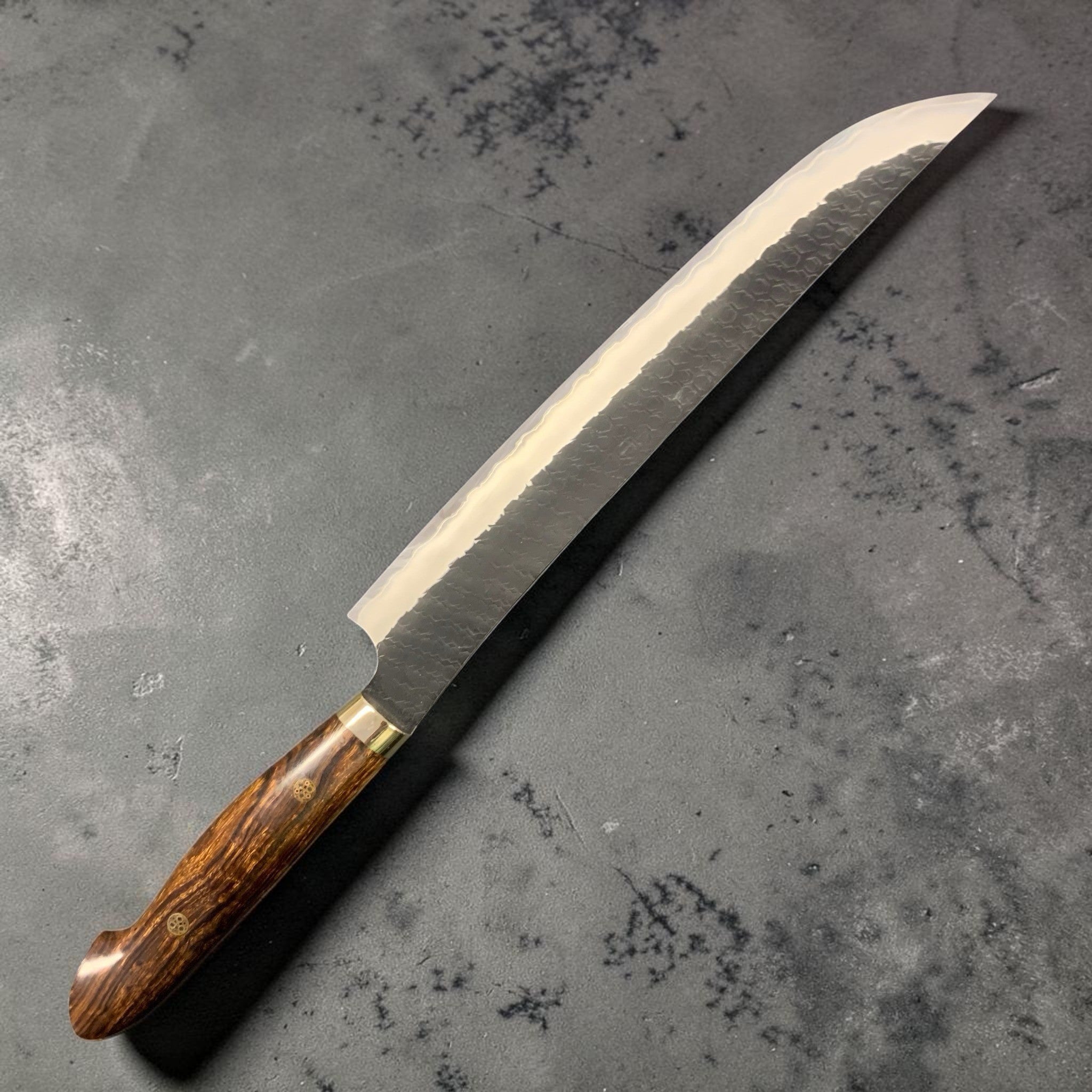 Nigara Hamono SG2 Kurouchi Tsuchime Sakimaru Sujihiki 270 mm, left-side profile highlighting the polished edge contrast against the dark kurouchi blade and ironwood Western handle.