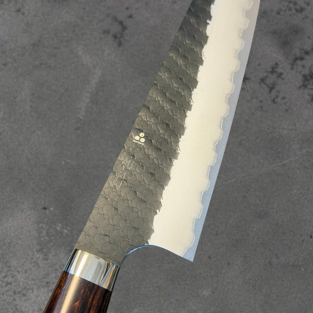 Close-up of the hammered Kurouchi finish, blade markings, and polished bevel on the Nigara SG2 Kurouchi Tsuchime 210mm gyuto with ironwood handle.