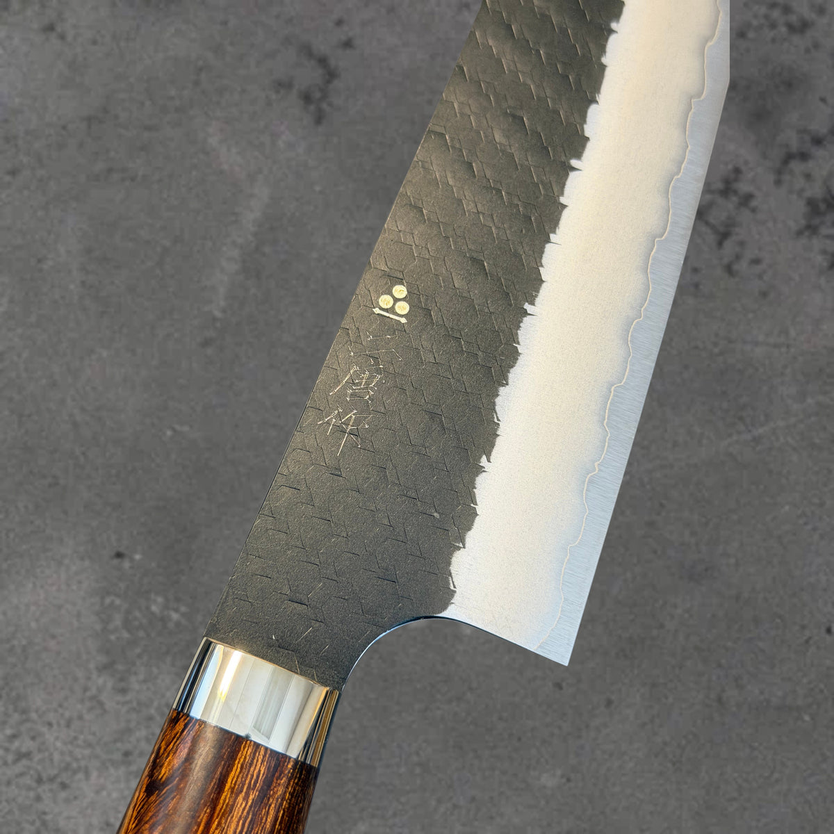 Close-up of the hammered Kurouchi finish, blade markings, and polished bevel on the Nigara SG2 Kurouchi Tsuchime 210mm kiritsuke with ironwood handle.