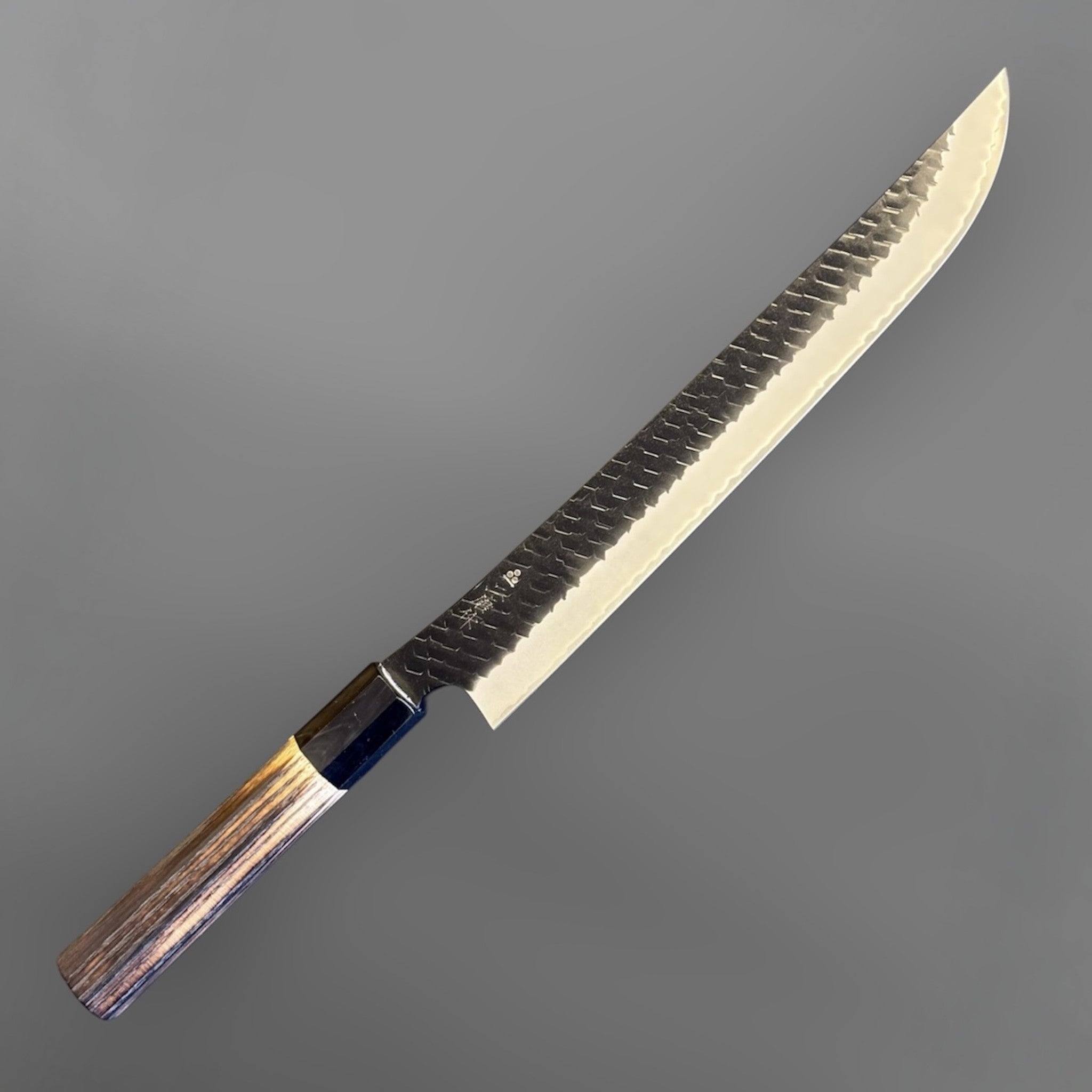 Nigara SG2 Tsuchime Kurouchi Sakimaru Sujihiki 270 mm, angled view highlighting the sweeping tip, dark hammered flats, and polished cutting edge.