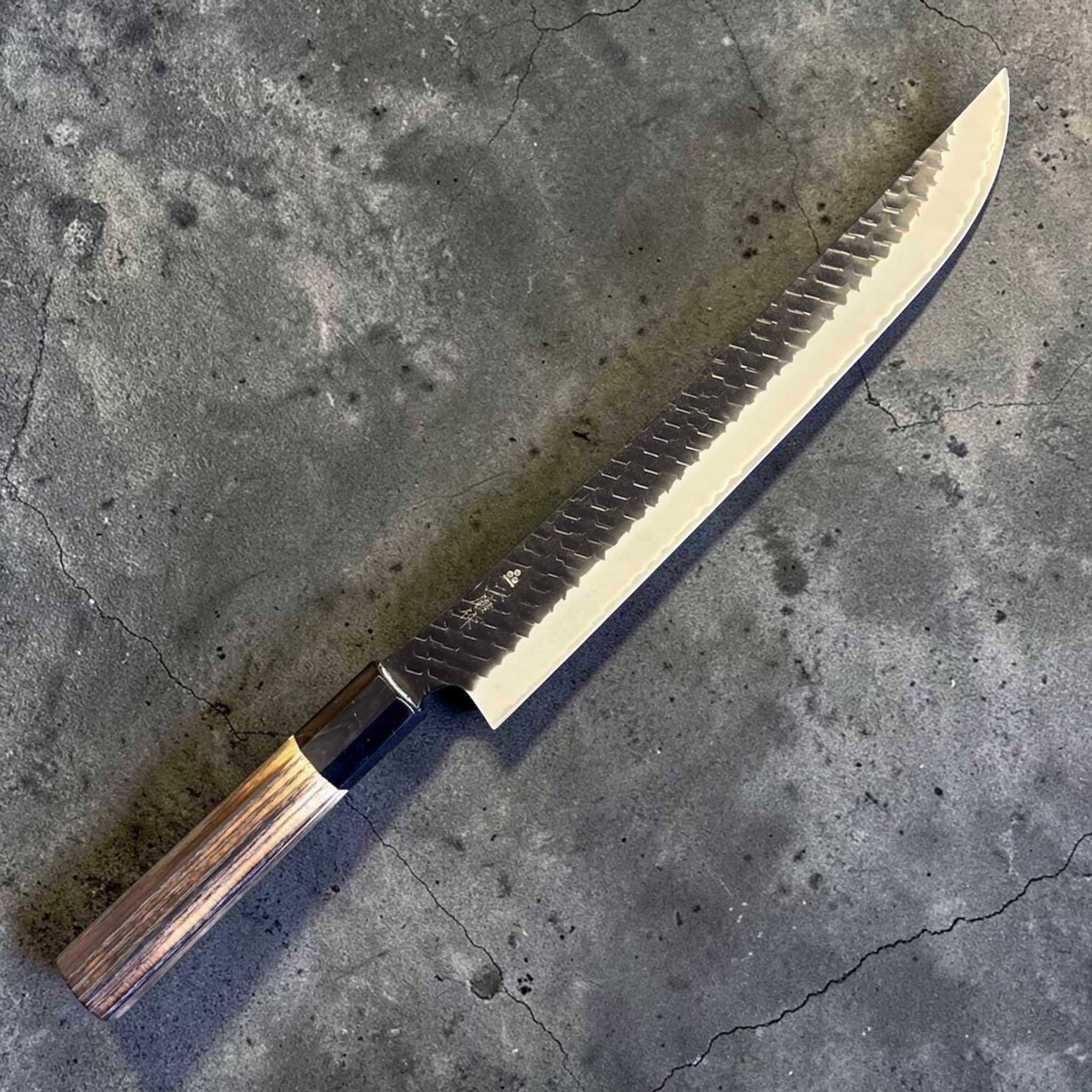 Nigara SG2 Tsuchime Kurouchi Sakimaru Sujihiki 270 mm, angled view highlighting the sweeping tip, dark hammered flats, and polished cutting edge.