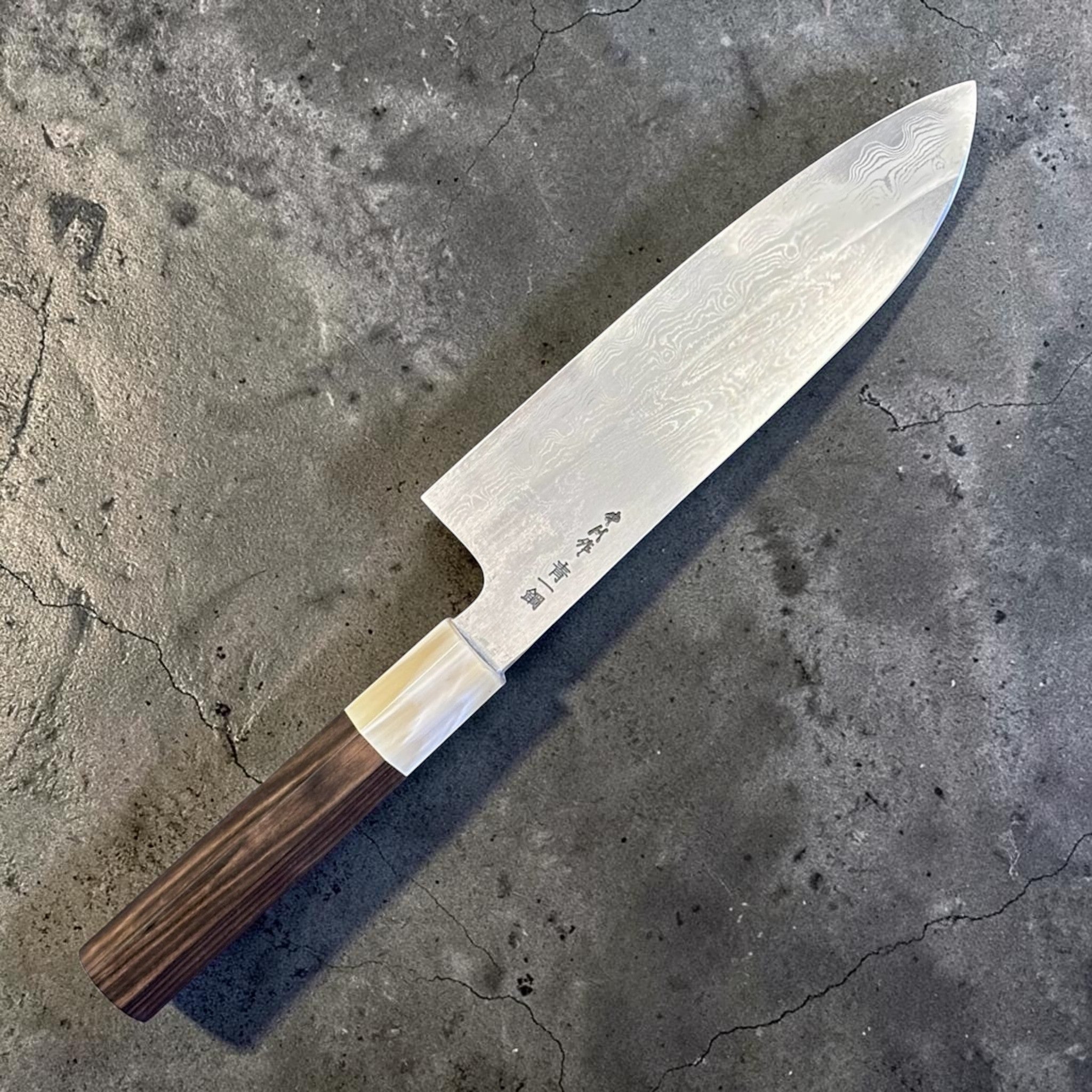 Satoshi Nakagawa Blue #1 Damascus Santoku with ebony and horn handle, reverse angled view highlighting kanji engraving, layered Damascus steel, and dark ebony wa handle with horn ferrule.