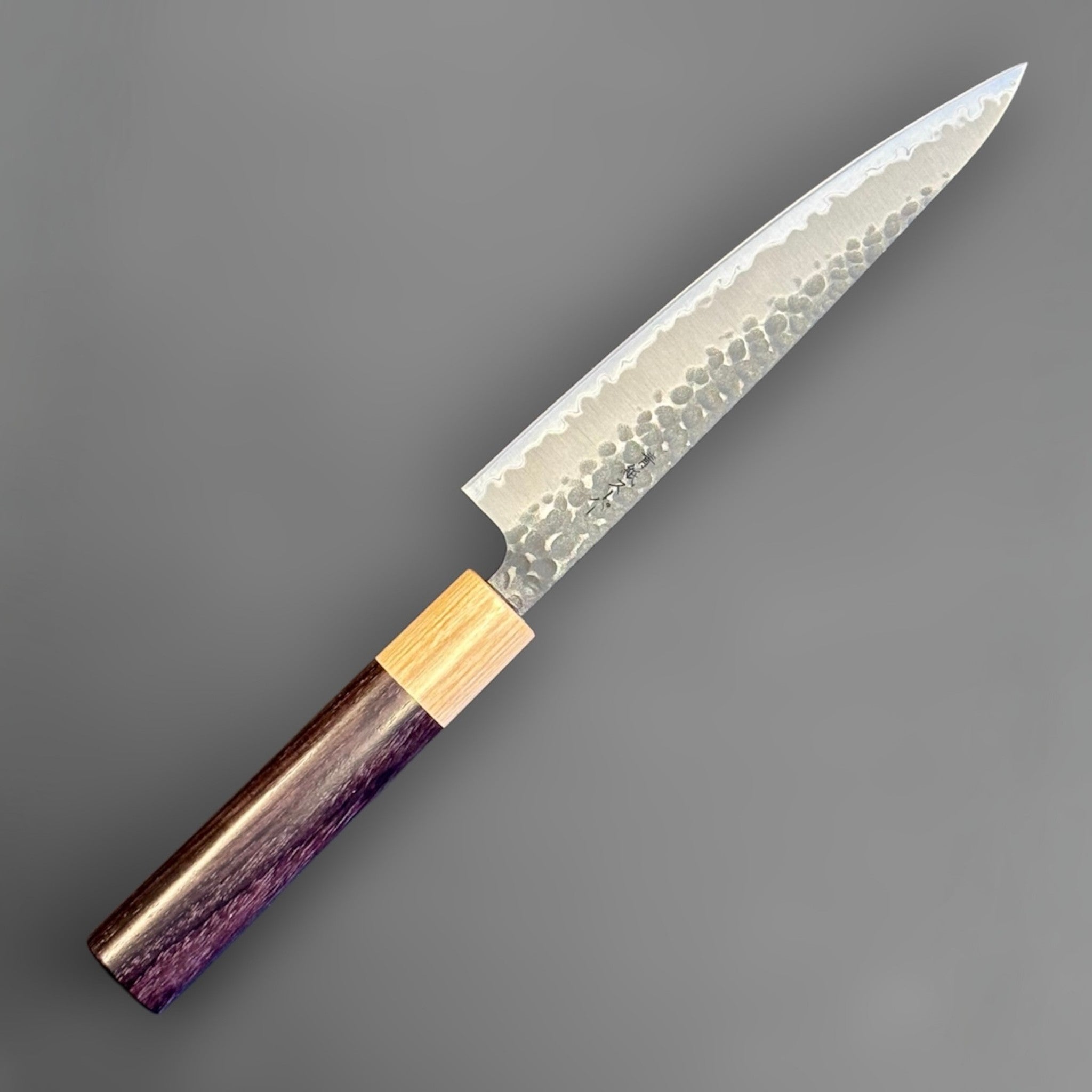Tsunehisa Aogami Super Tsuchime Kuro Petty 150 mm with morado handle, angled reverse-side view highlighting the hammered blade pattern, migaki core line, and slim wa handle.