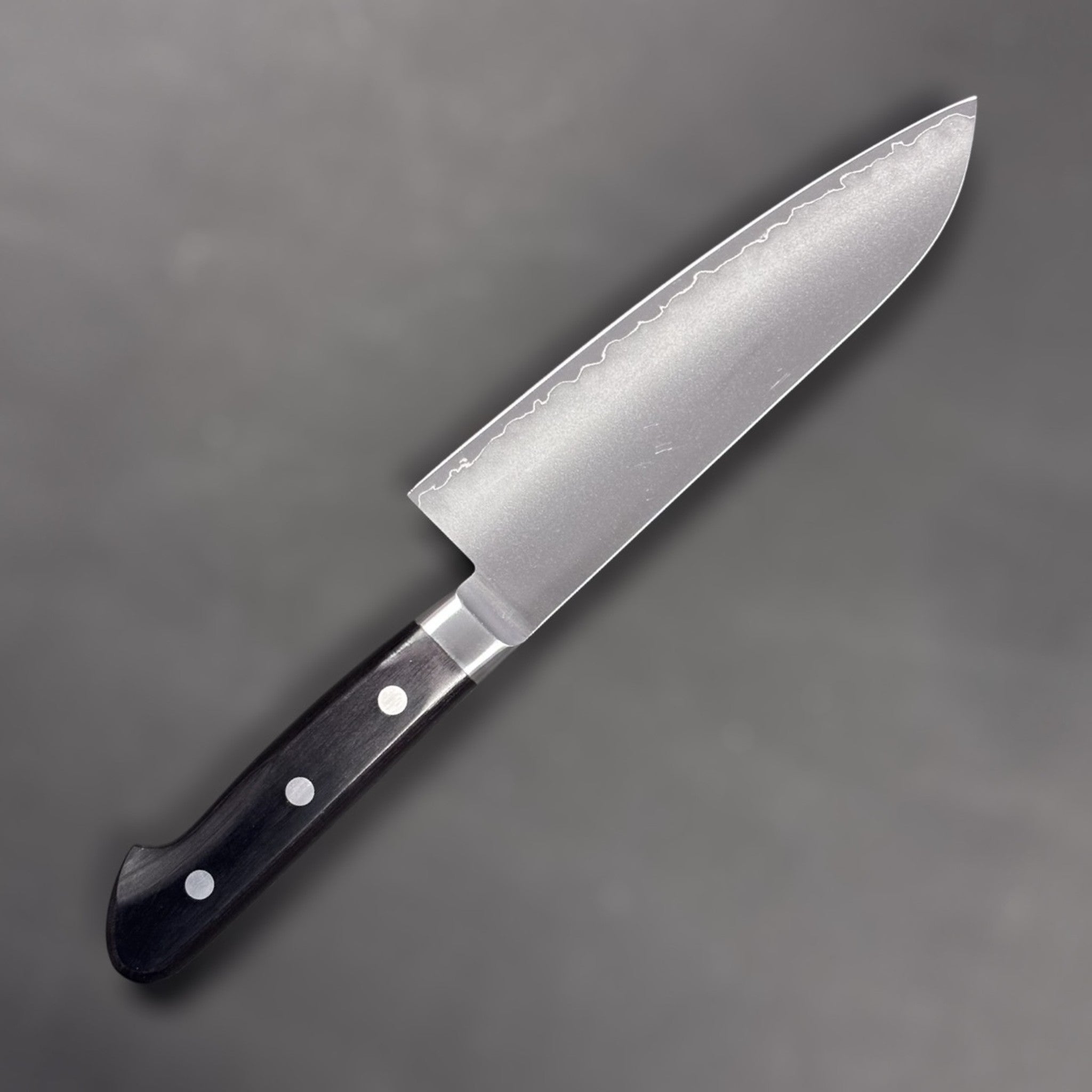 Tsunehisa VG-1 stainless steel Santoku 165 mm with black Western handle, left side blade face highlighting the soft cladding line.