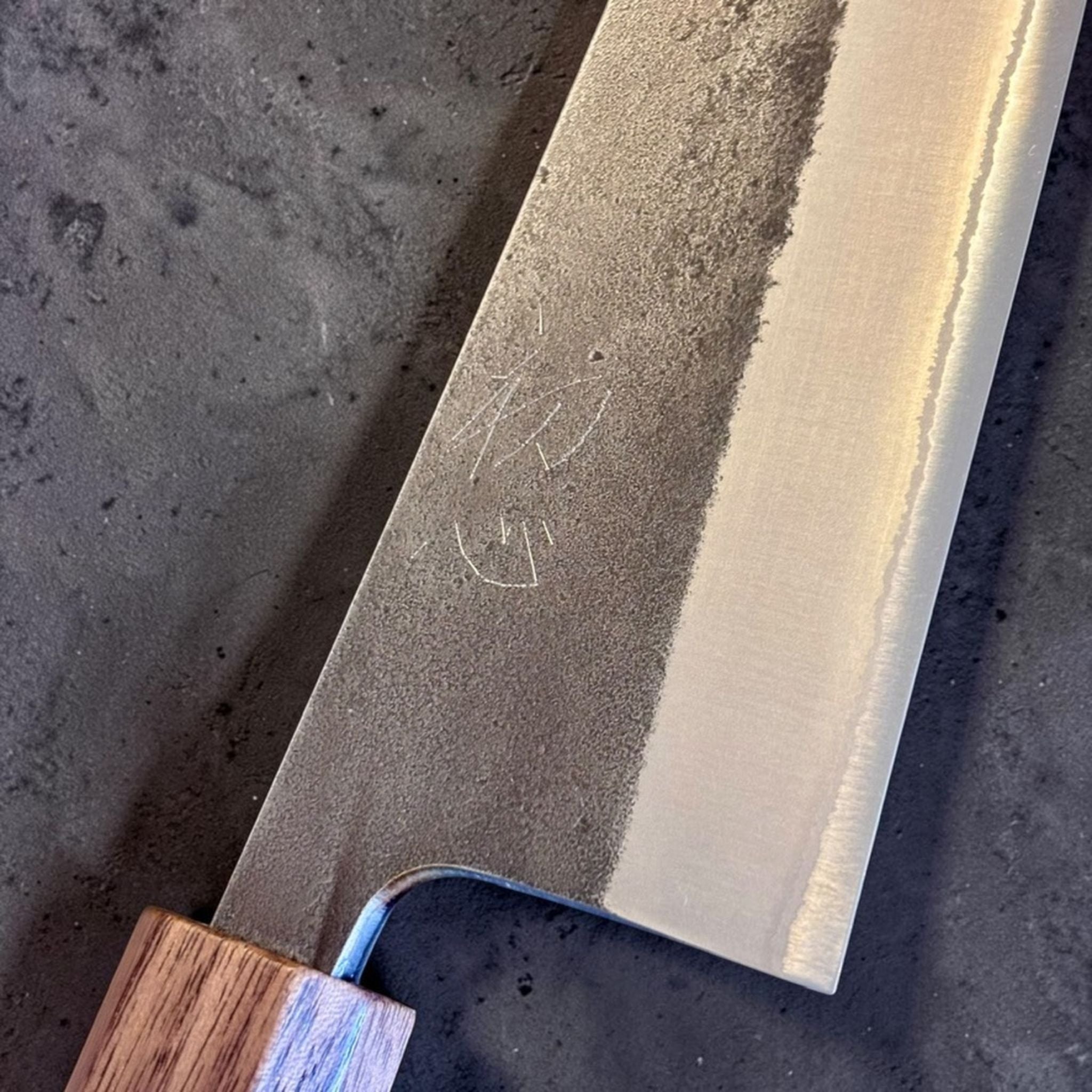Yoshikane Nashiji Kurouchi Santoku with Sanjo walnut handle, close-up of the nashiji kurouchi blade texture and maker engraving.